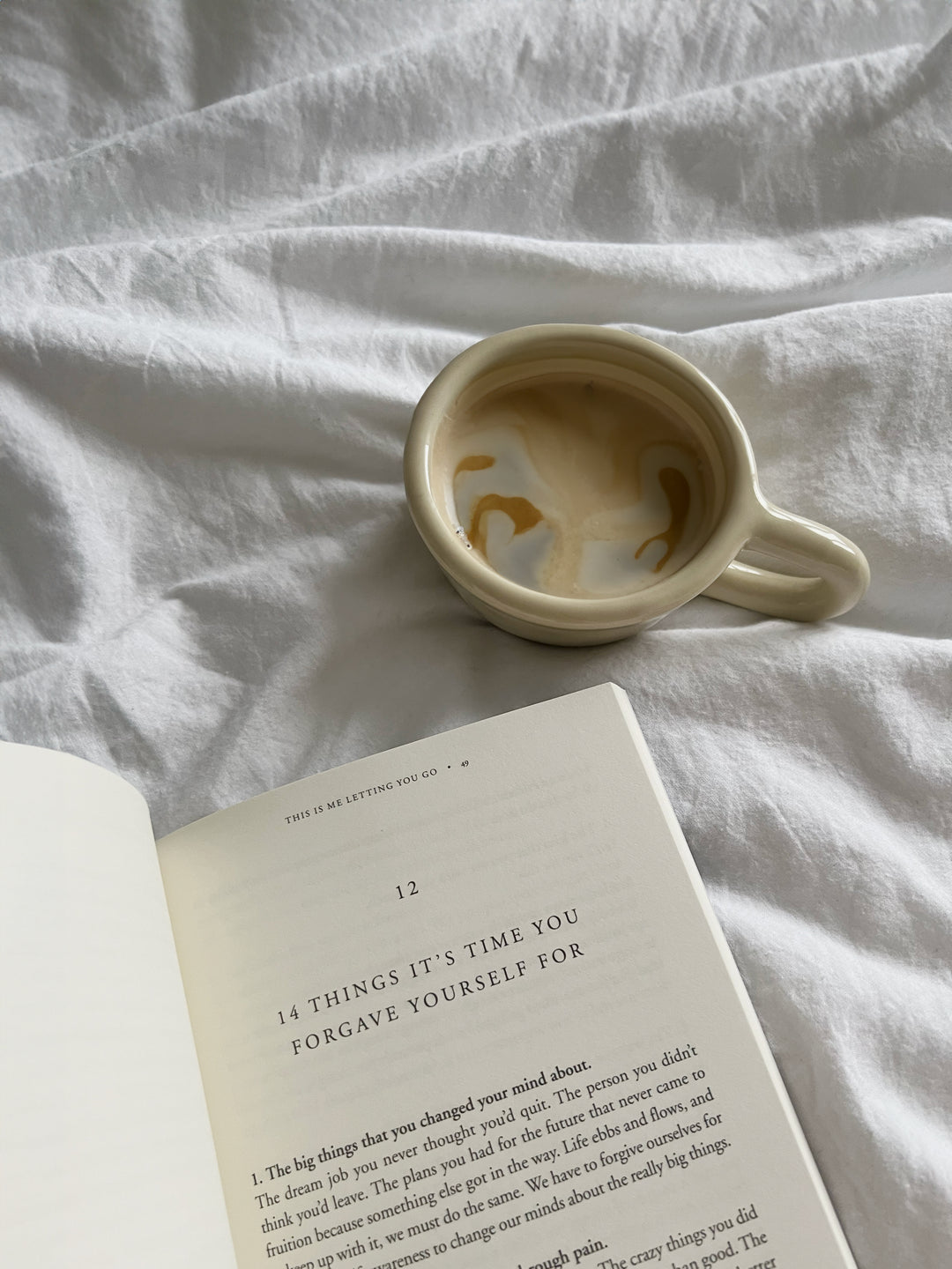 Coffee cup beside the book open to a page on forgiving yourself, on white bedding