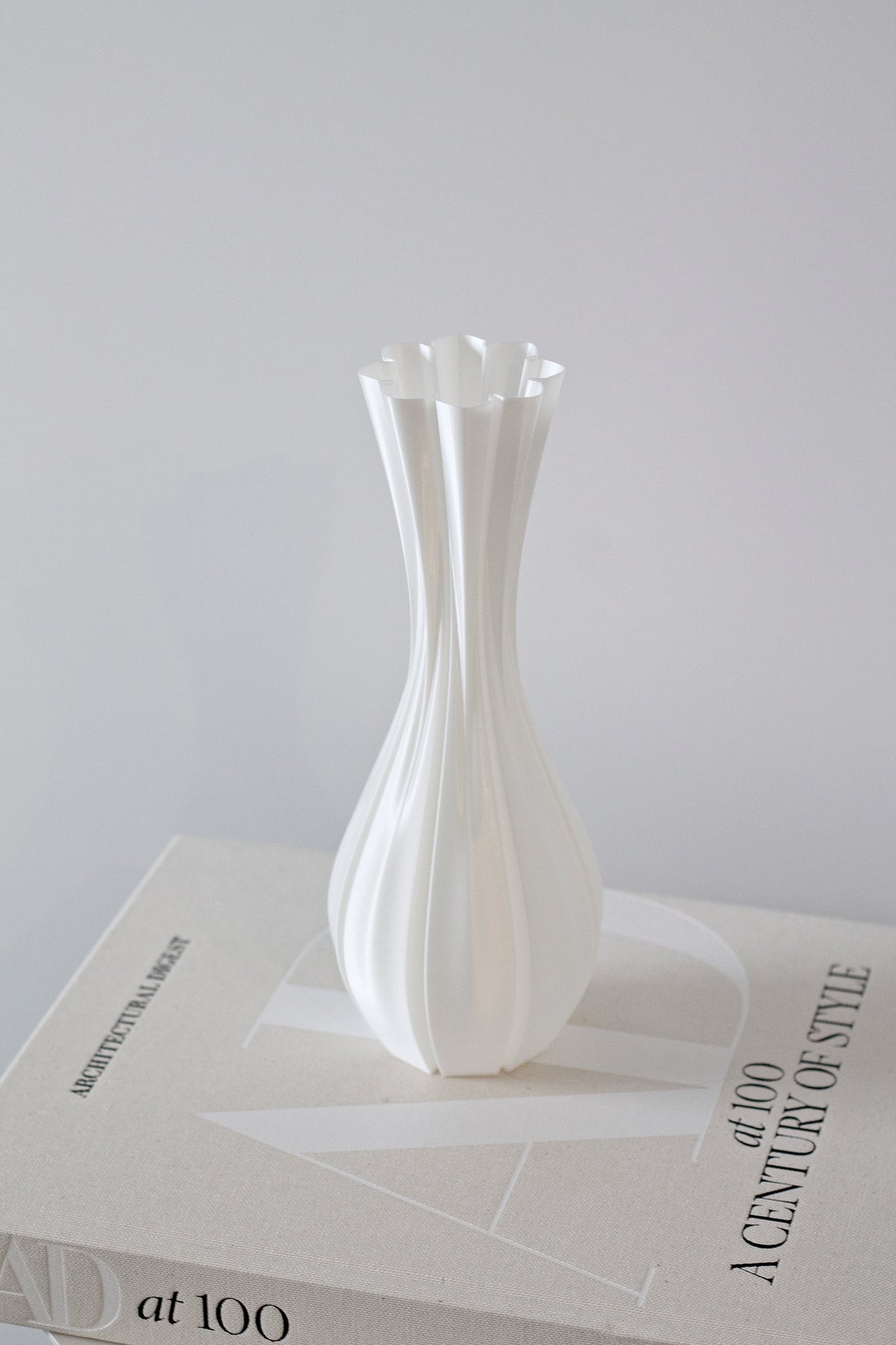 Fluted white vase standing on stacked books against a pale grey wall, full height in view