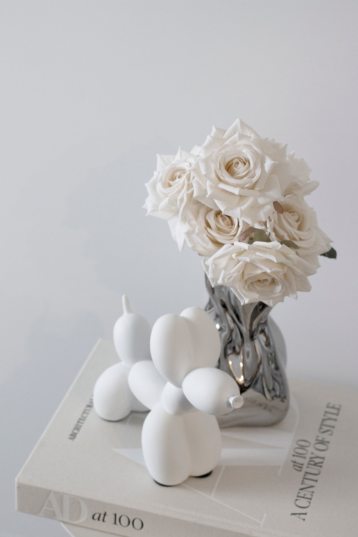Champagne roses in a silver vase beside a white balloon dog on coffee table books