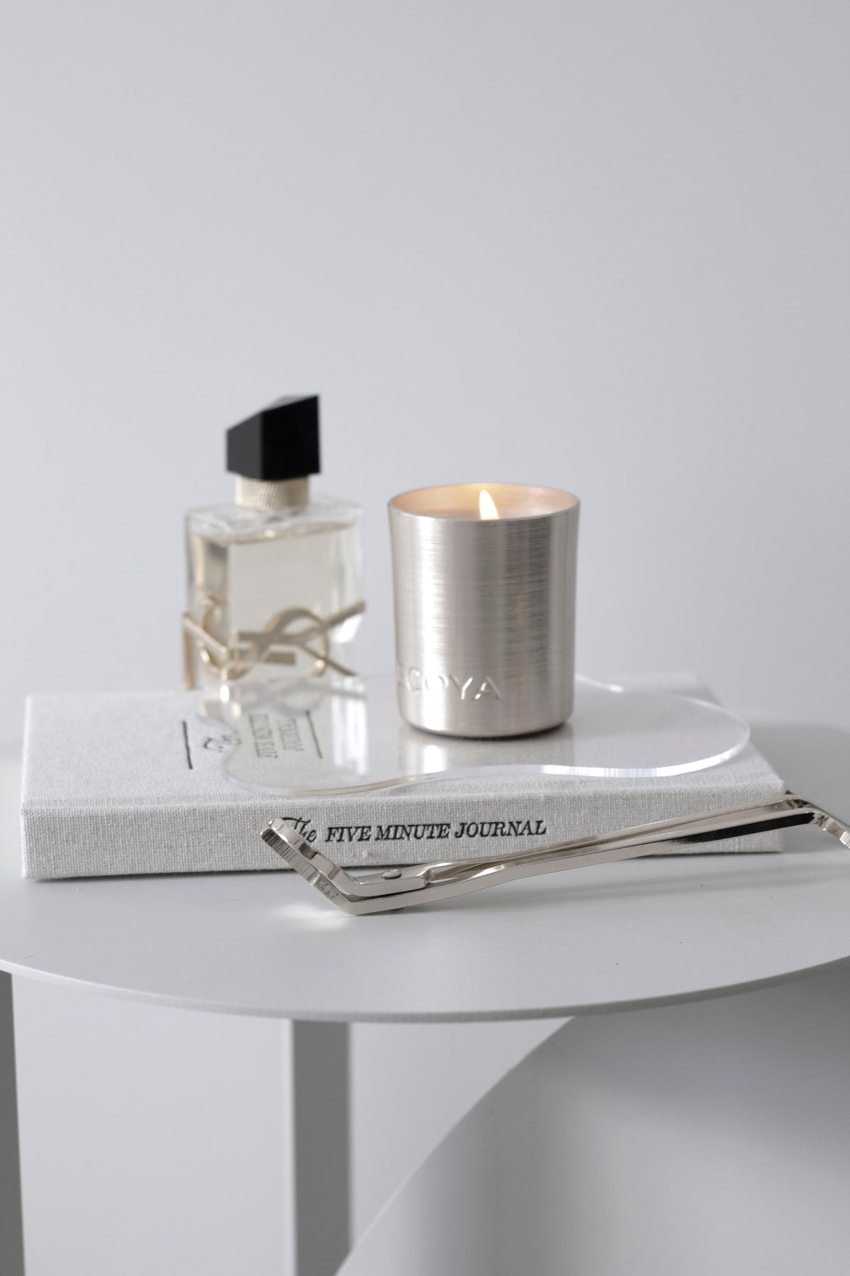 Calming flatlay featuring a lit ECOYA candle, a bottle of perfume, and The Five Minute Journal stacked on a round white side table with a silver wick trimmer
