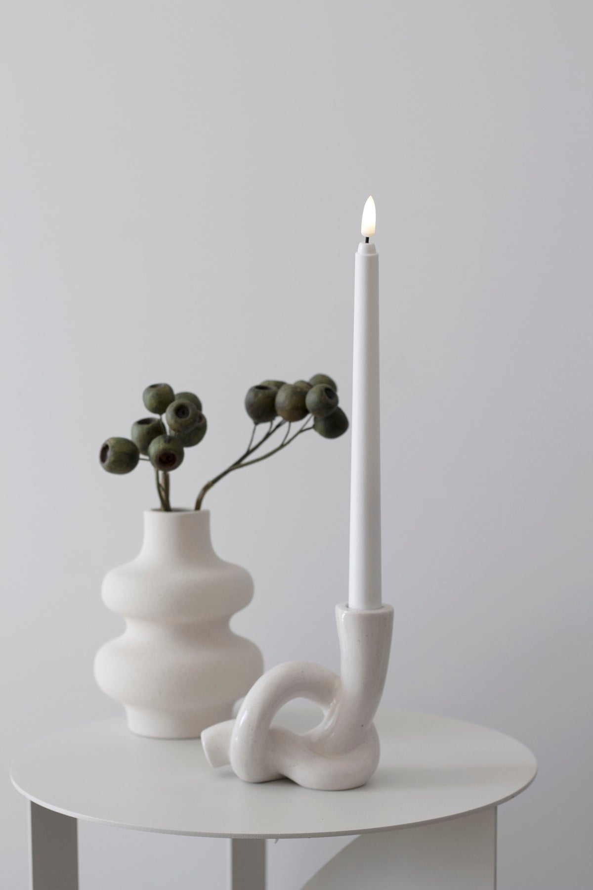 Lit taper in the knot holder on a pale side table, a tiered vase of dried gum nuts alongside