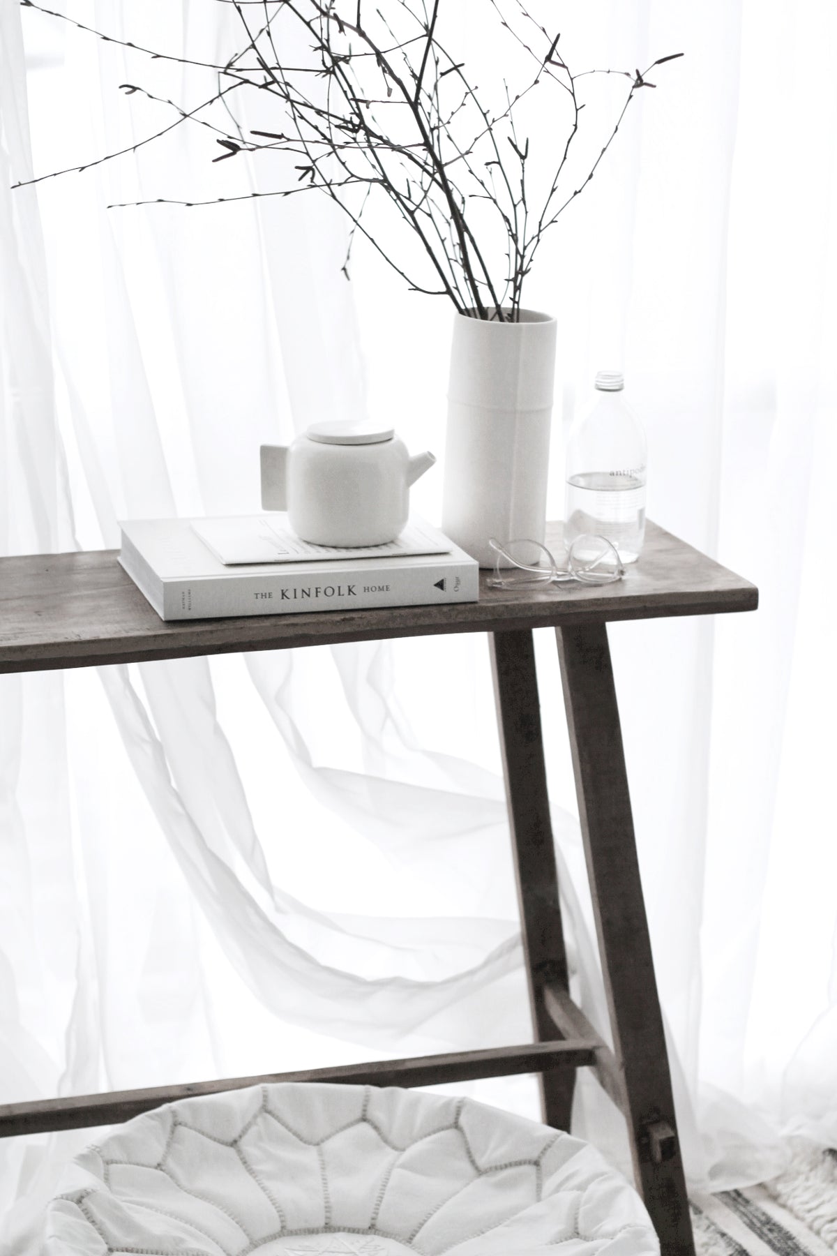 Recycled teak console with a tall white vase of bare branches and books, sheer curtain behind