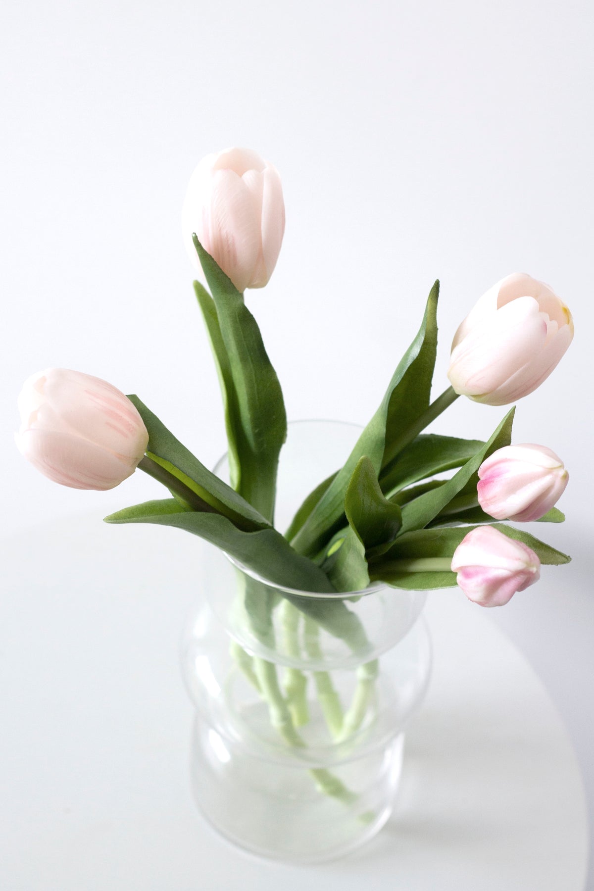 Pale pink faux tulips arranged low in a clear glass vase against a white wall