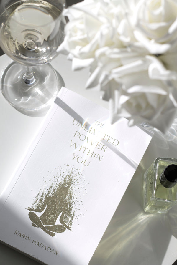 Flatlay of The Unlimited Power Within You by Karin Hadadan with white roses, wine glass, and perfume bottle on a glossy white surface.