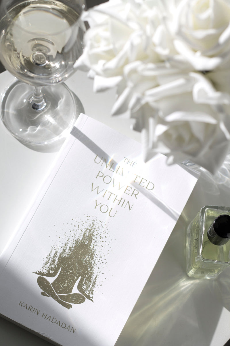 Flatlay of The Unlimited Power Within You by Karin Hadadan with white roses, wine glass, and perfume bottle on a glossy white surface.