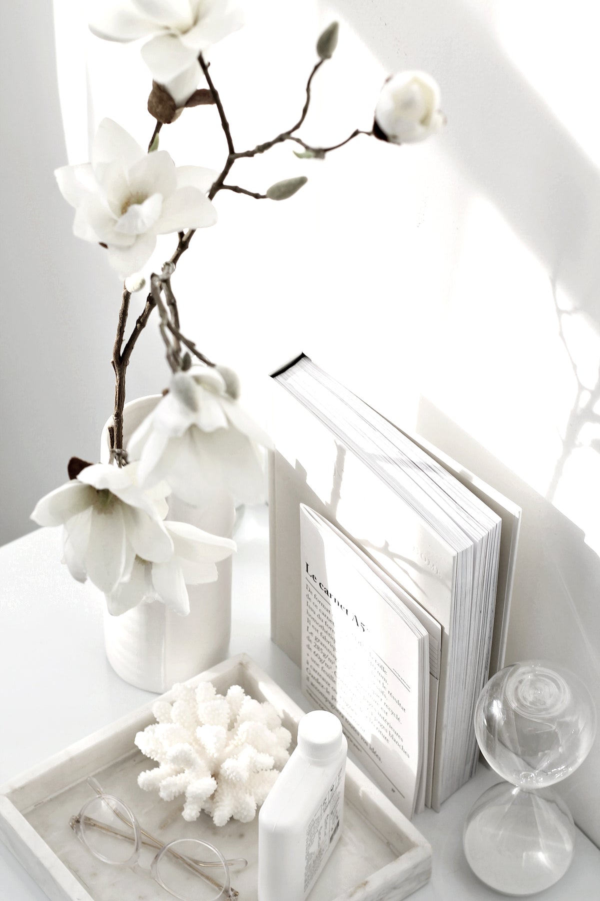 Hourglass on a white tray with magnolia stems, a book and a coral piece in bright light