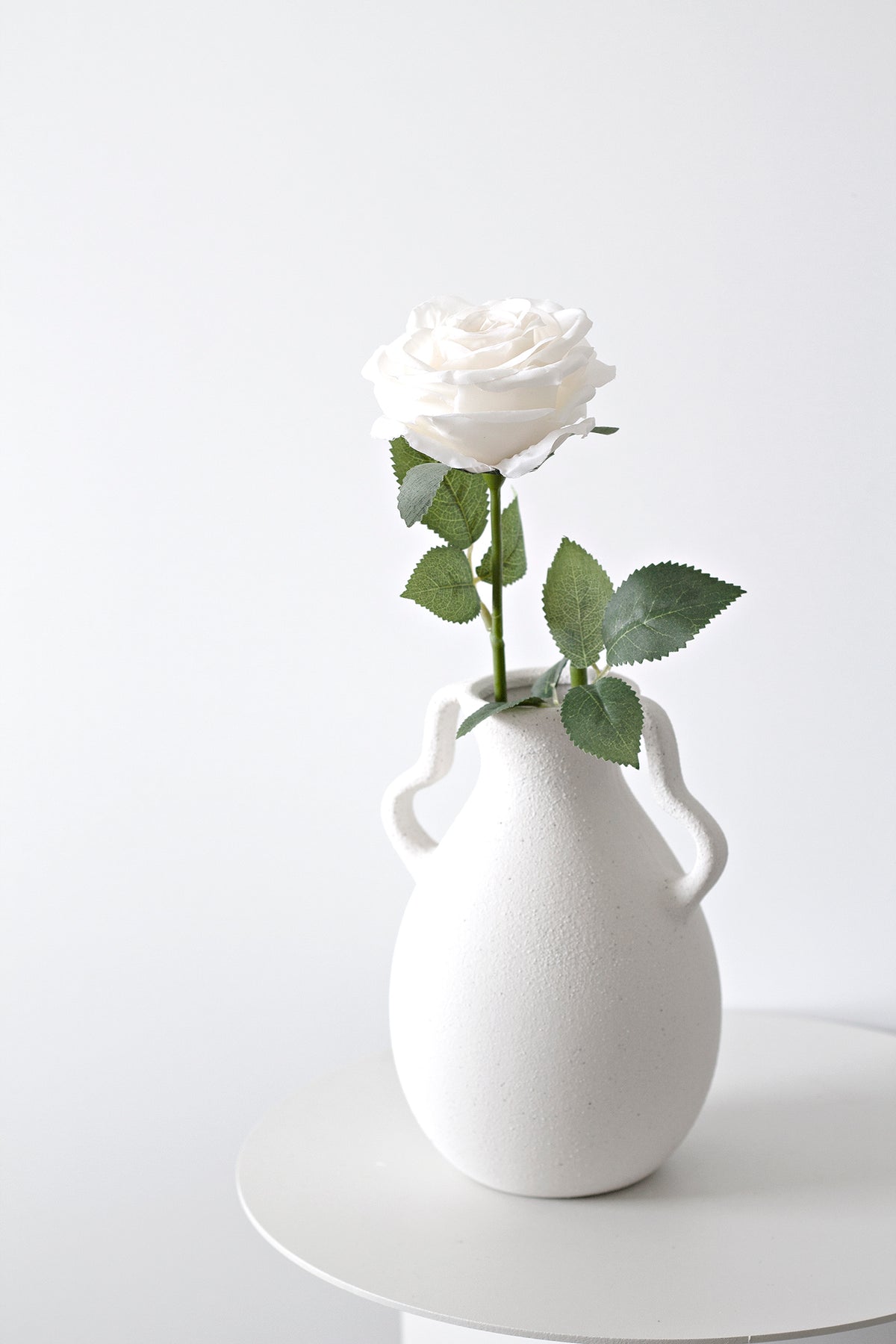 Cream rose in a small white handled vase on a round pedestal
