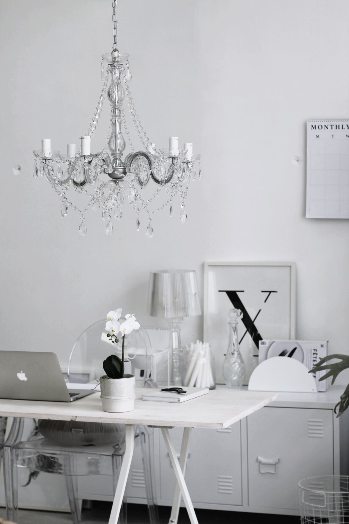 Clear acrylic chair at a white desk below a crystal chandelier, with a framed letter print