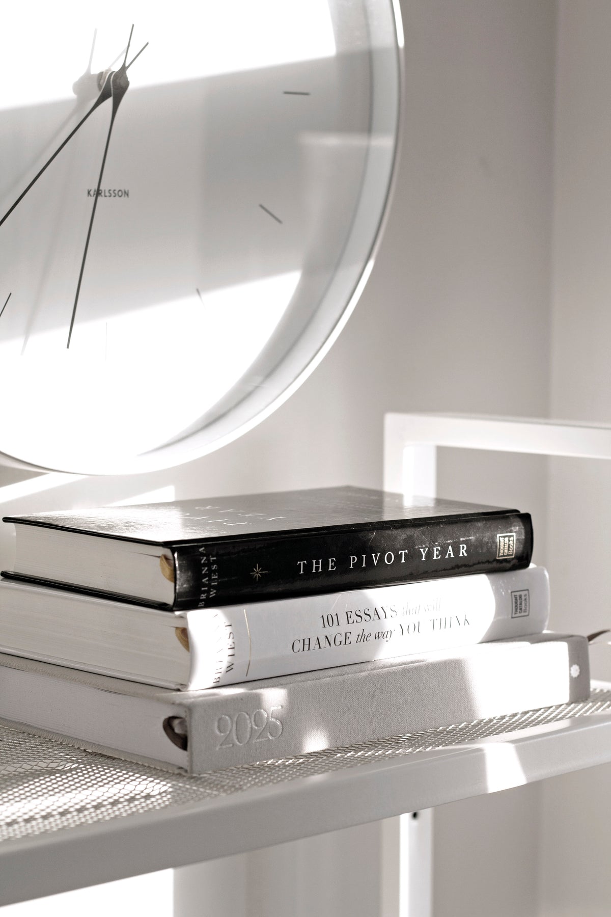 Books stacked on a white mesh shelf beneath a round wall clock, a picture frame leaning behind
