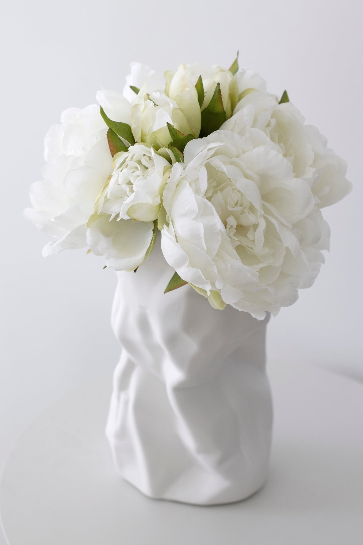 White crumpled vase filled with white peonies on a pale surface