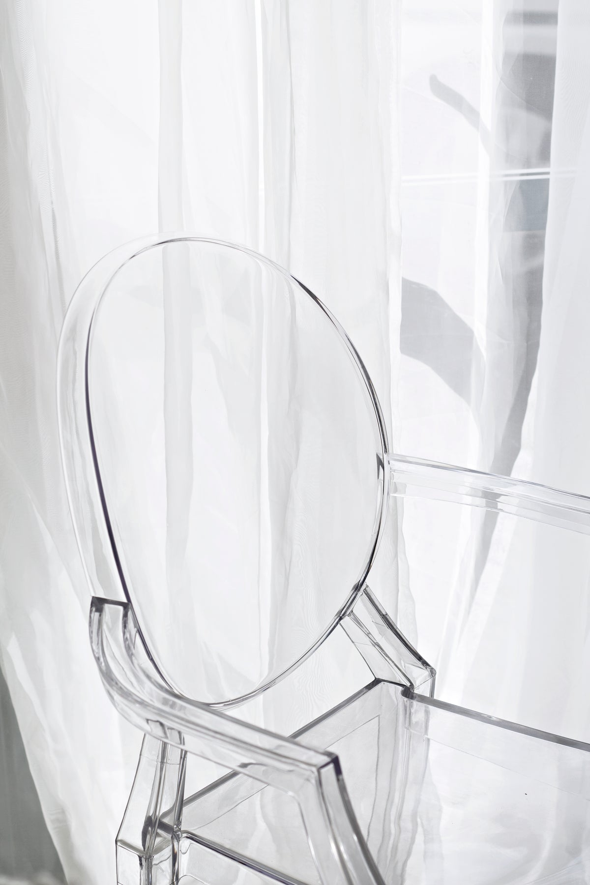 Oval back of the clear acrylic chair against sheer white curtains in soft daylight
