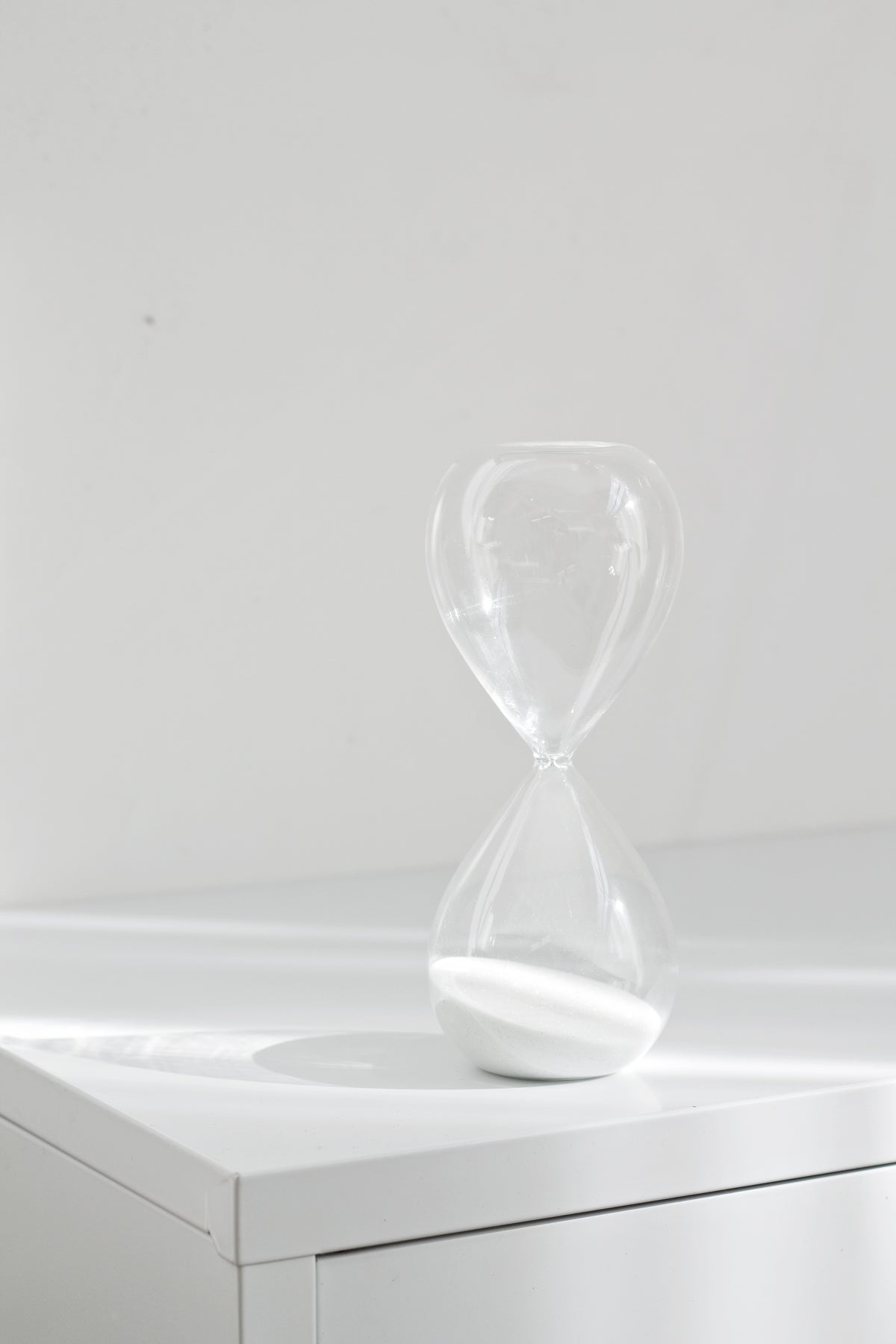 Frameless hourglass standing alone on a white cabinet, sand collected in the lower bulb