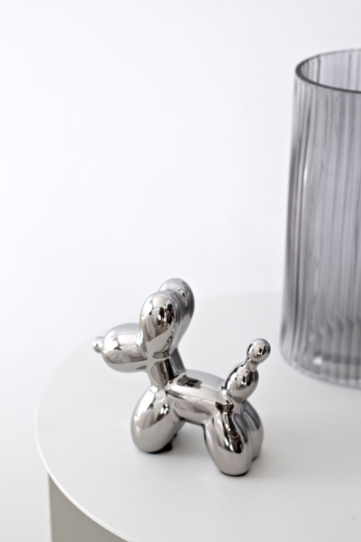 Small silver balloon dog sculpture beside a ribbed glass vase on a white table