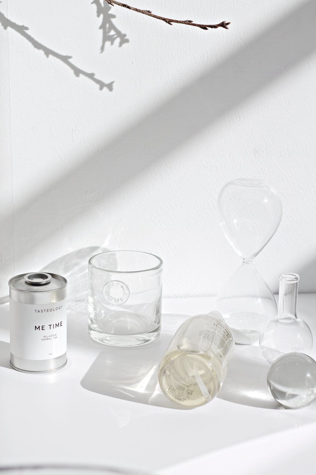 Hourglass upright beside a tumbler, a small flask and a tin on a sunlit white surface
