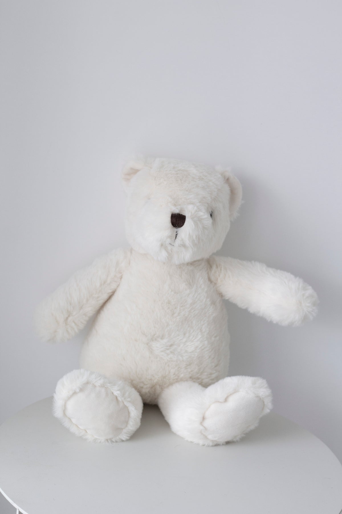 Warm White Flopsy Bear sitting on a white table against a pale grey wall, arms out
