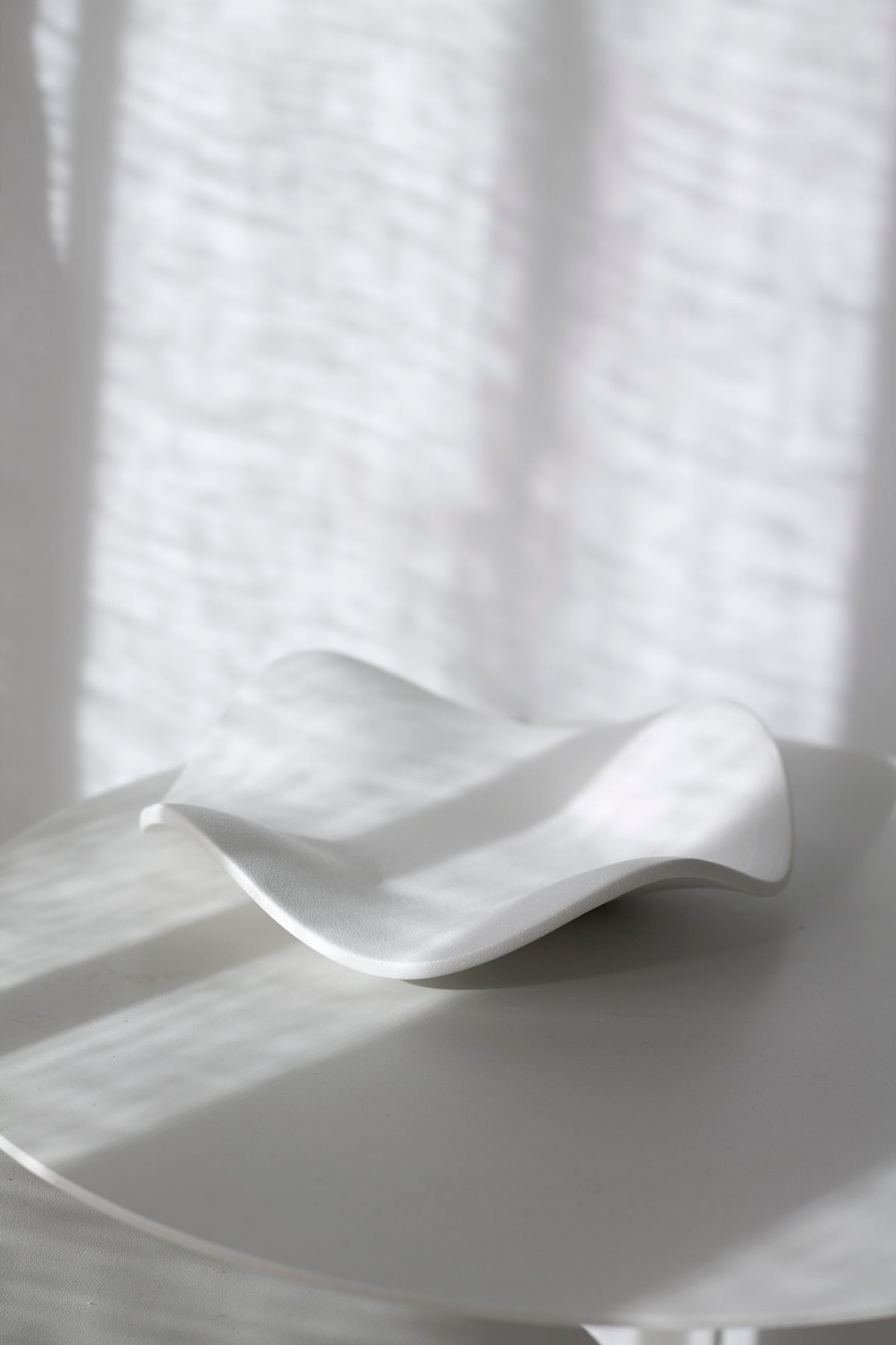 White wave tray on a round white table, the rippled rim lit softly against a sheer curtain