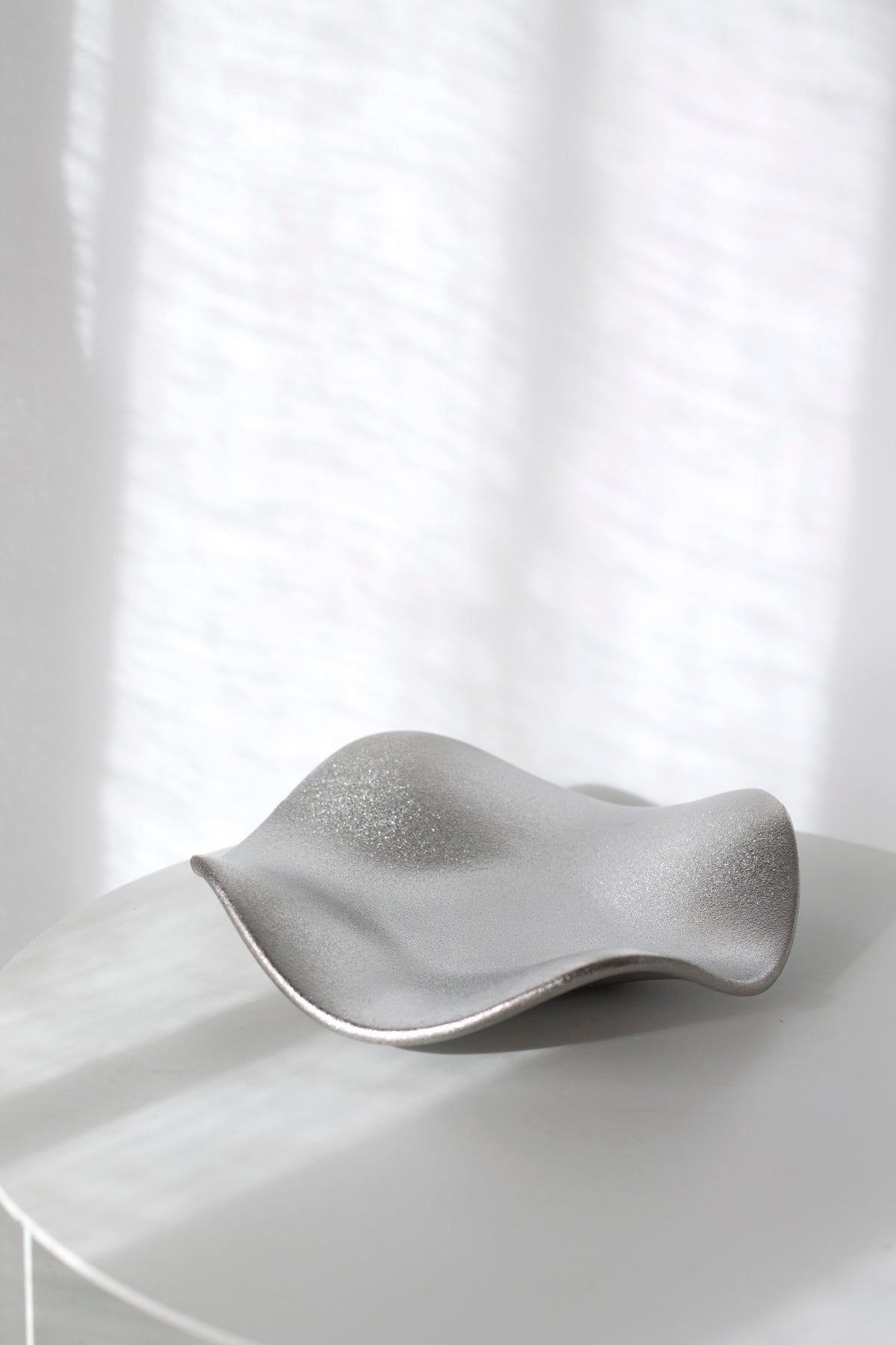 Silver wave tray with a finely speckled metallic finish on a round white side table by a sheer curtain