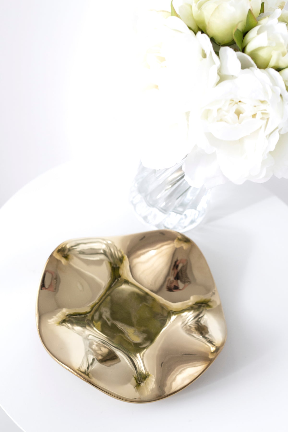 Polished gold wave tray on a white table with white blooms in a fluted clear glass vase behind