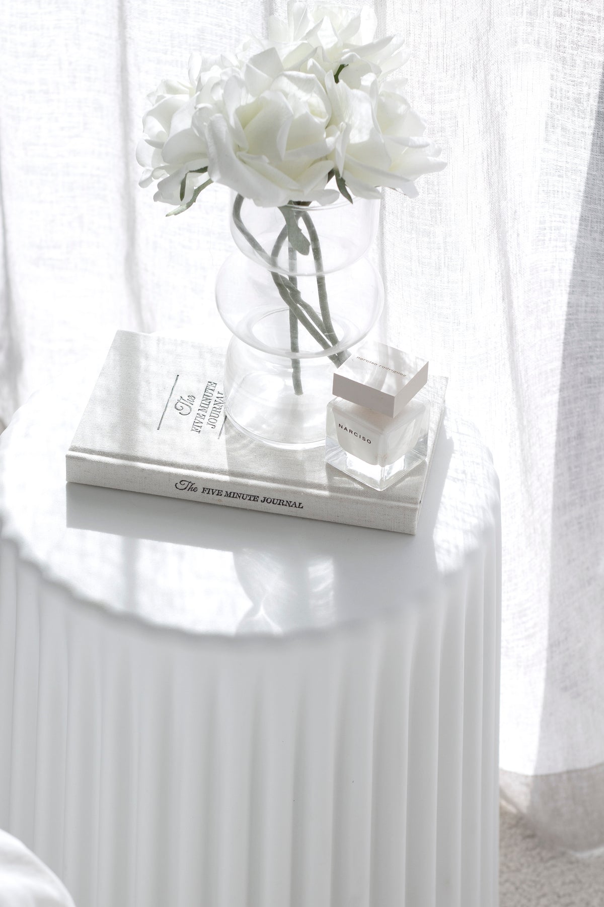 White ribbed stool holding a glass vase of white roses on books, bright light through the curtain