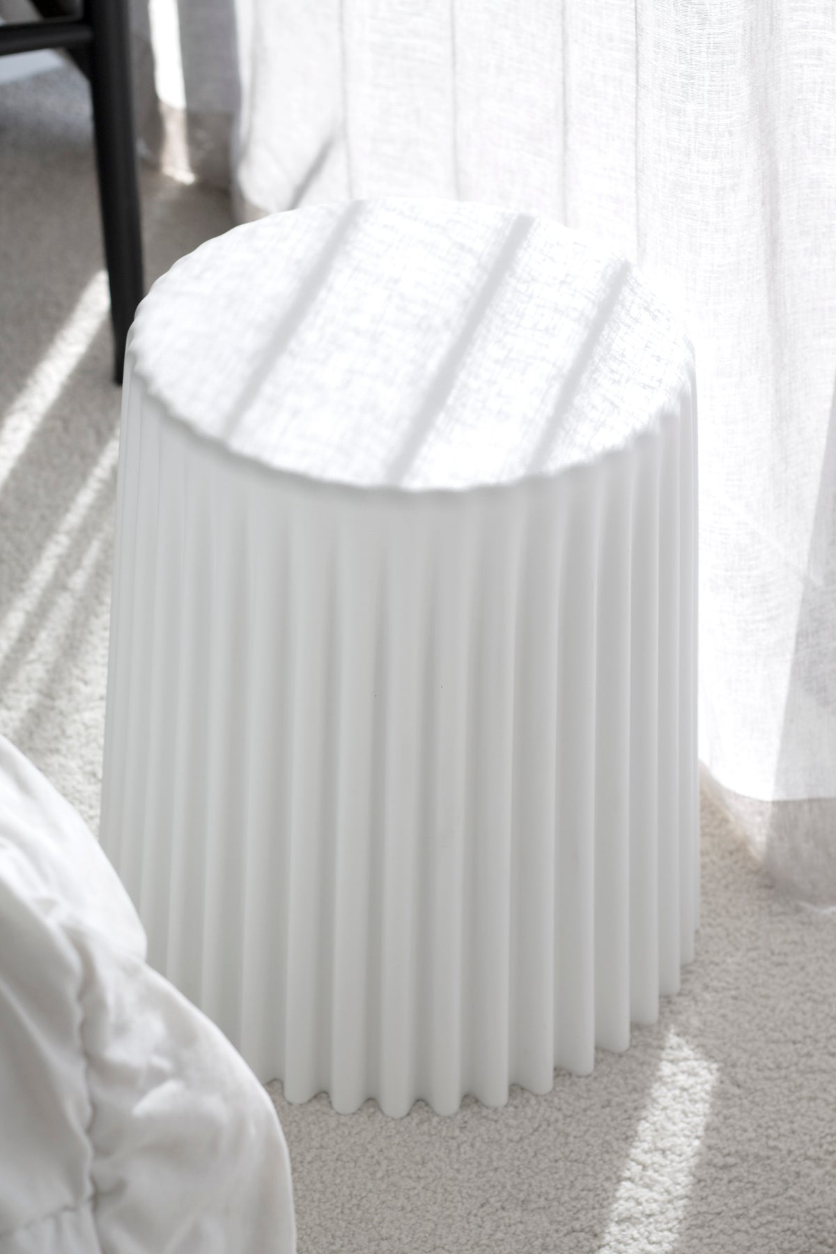 White ribbed stool on carpet beside a bed, strong sunlight striping the sheer curtain behind