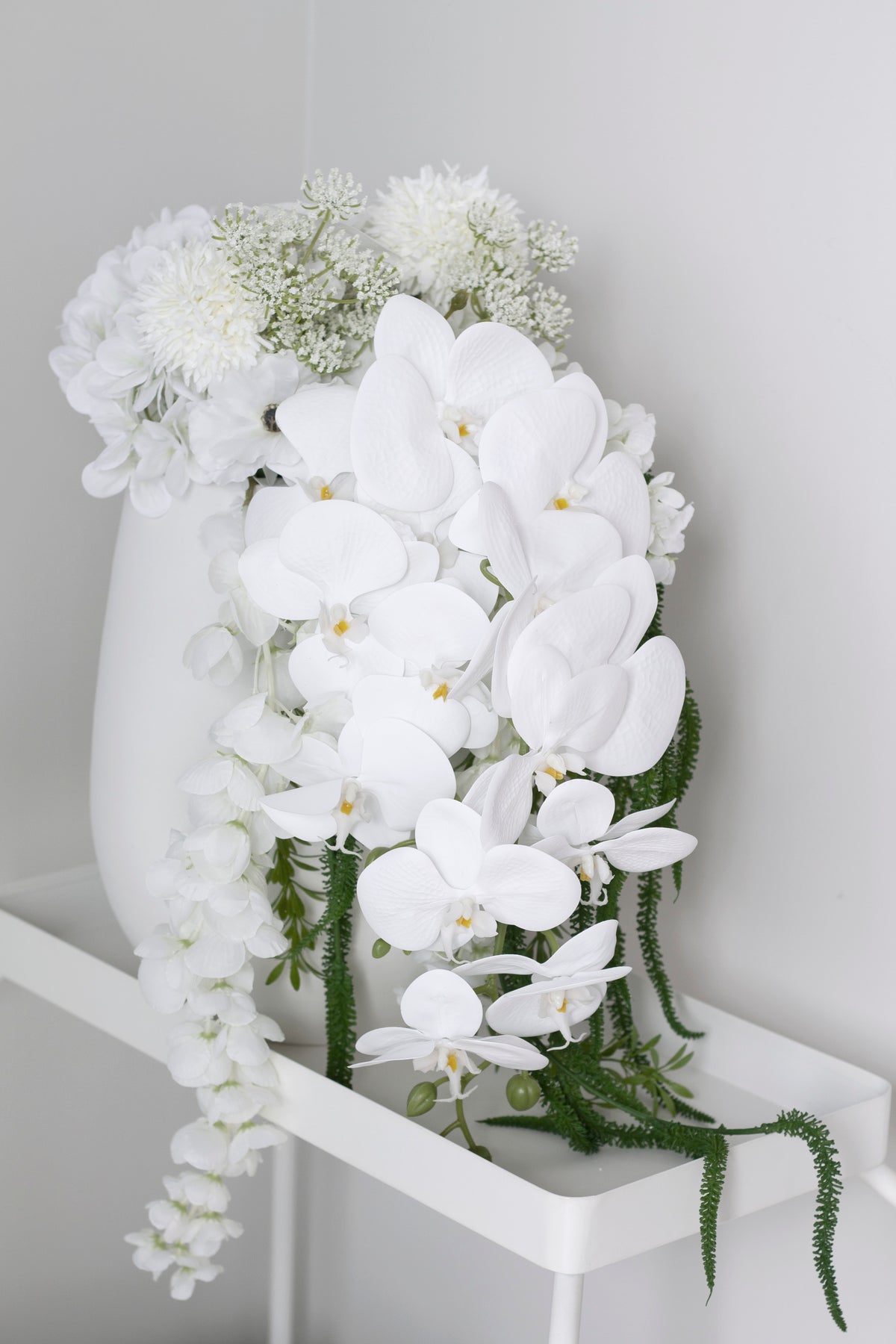The Estella bouquet in a tall white vase on a pale plinth, trailing stems falling to one side