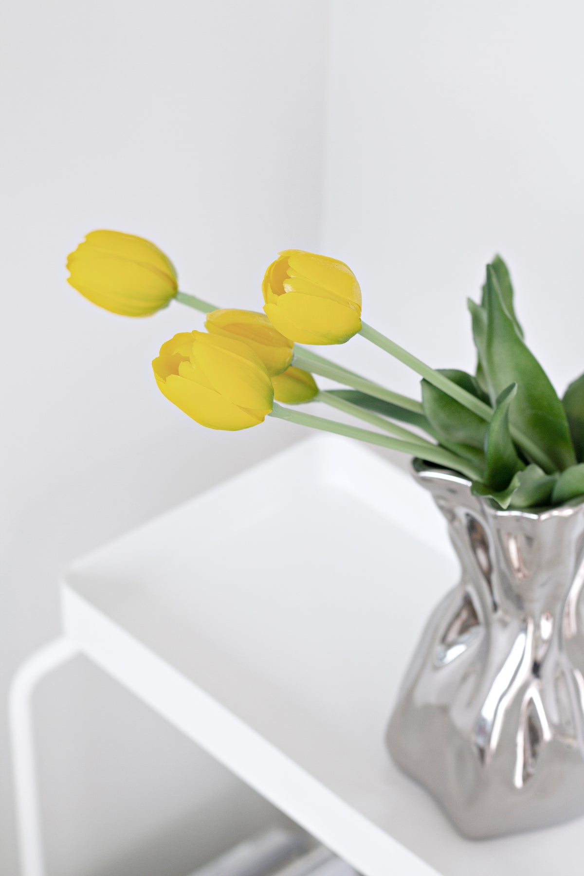 Yellow faux tulips in a silver crumpled vase on the corner of a white side table