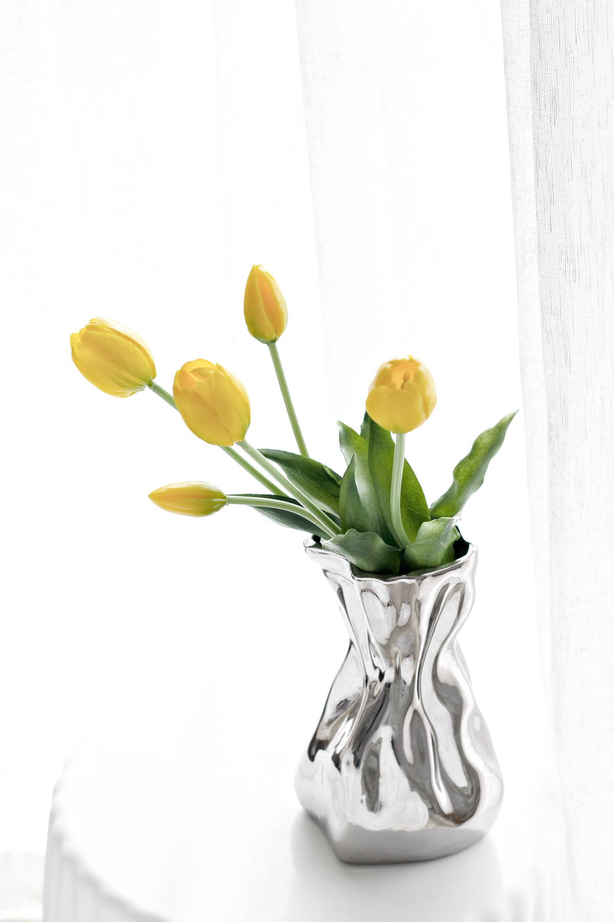 Yellow faux tulips arranged in a silver crumpled vase on a white side table by a sheer curtain