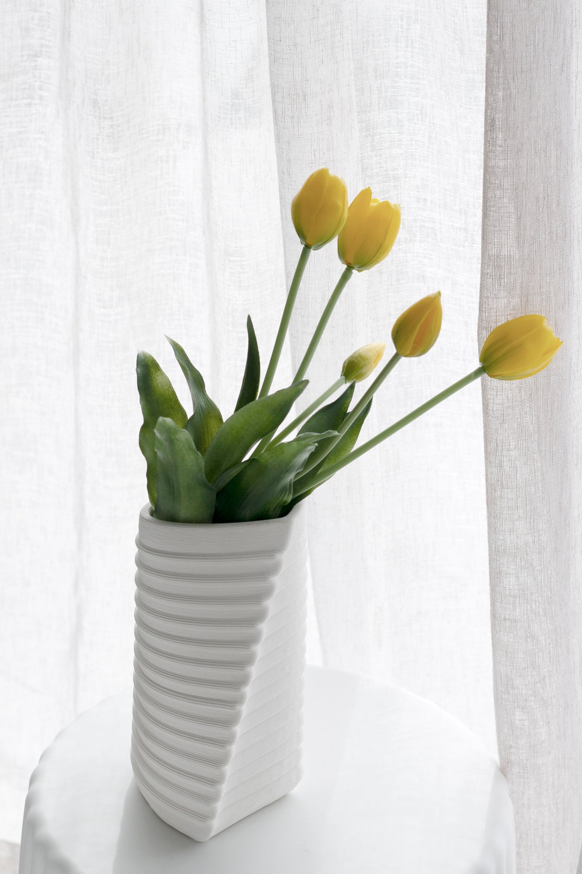 Yellow faux tulips in a ribbed white ceramic vase on a round side table by a sheer curtain
