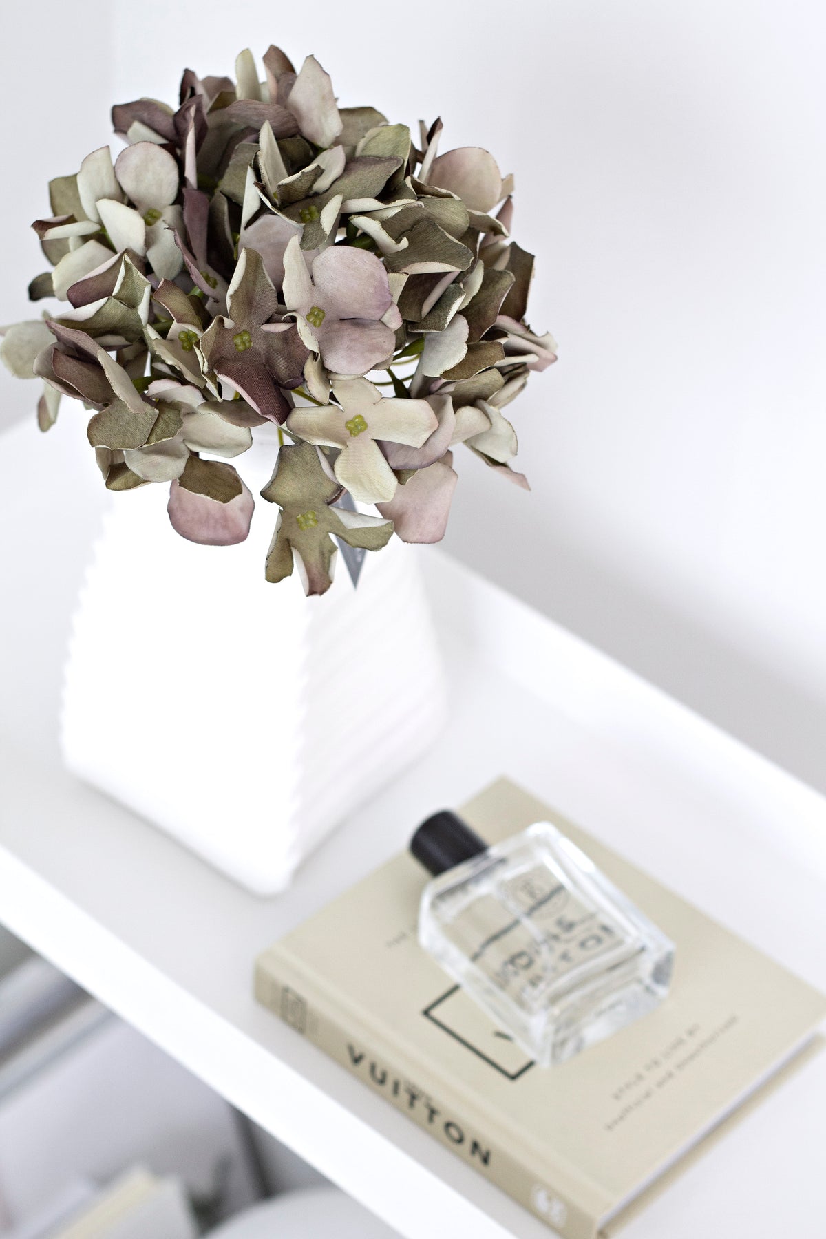 Green faux country hydrangea in a white vase on a shelf beside a book and perfume bottle