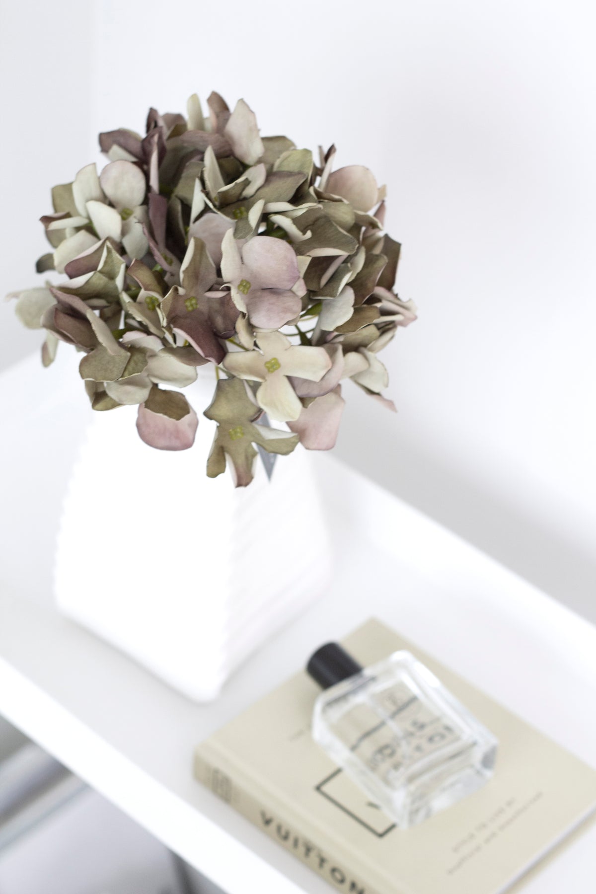 Green faux hydrangea in a white vase on a shelf with a book and perfume bottle