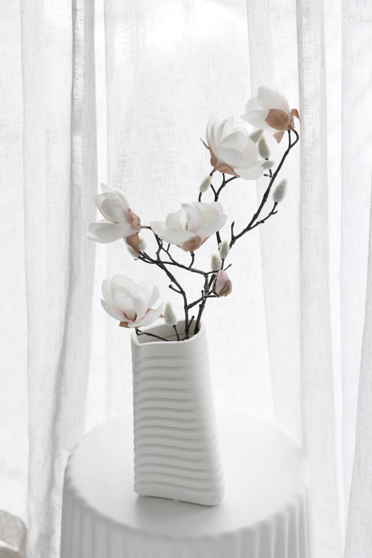 Faux magnolia blooms arching from a ribbed white vase by a window
