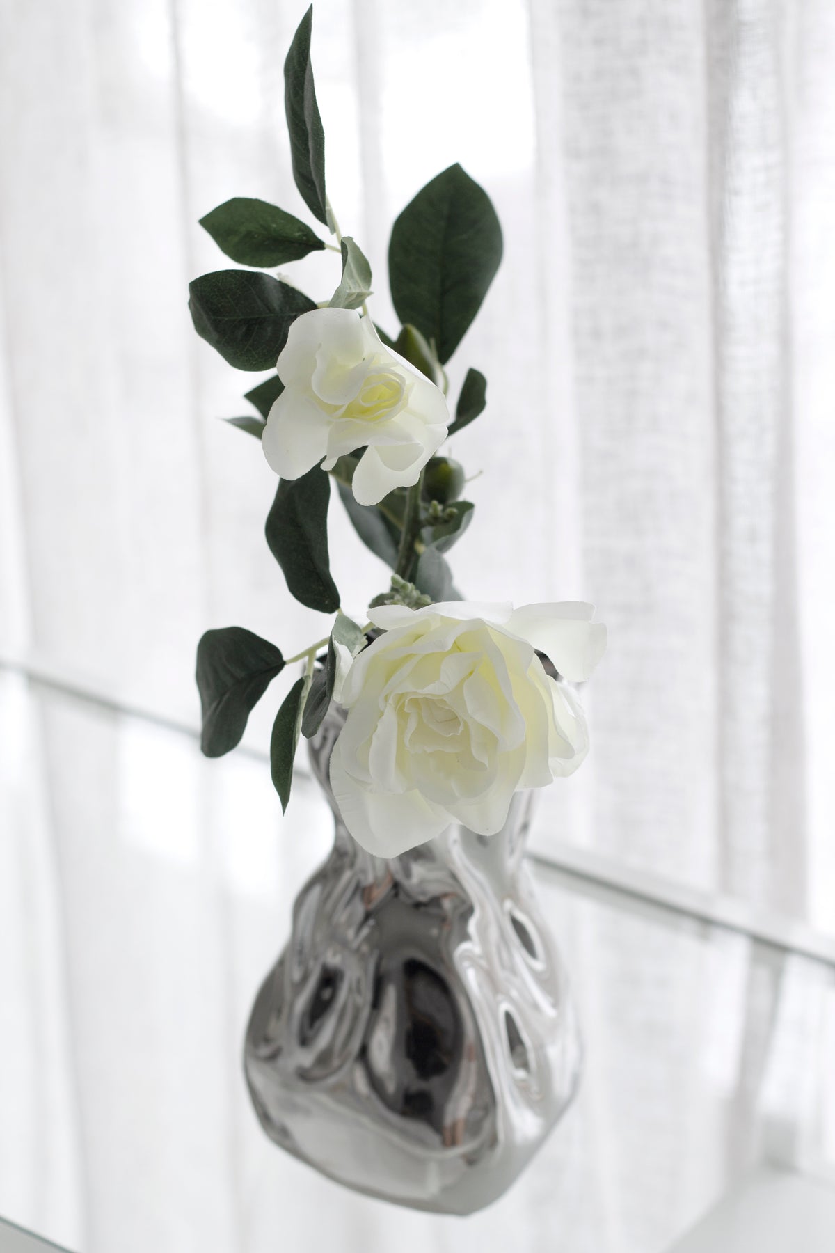 Gardenia stem in a textured silver vase against a sheer curtain