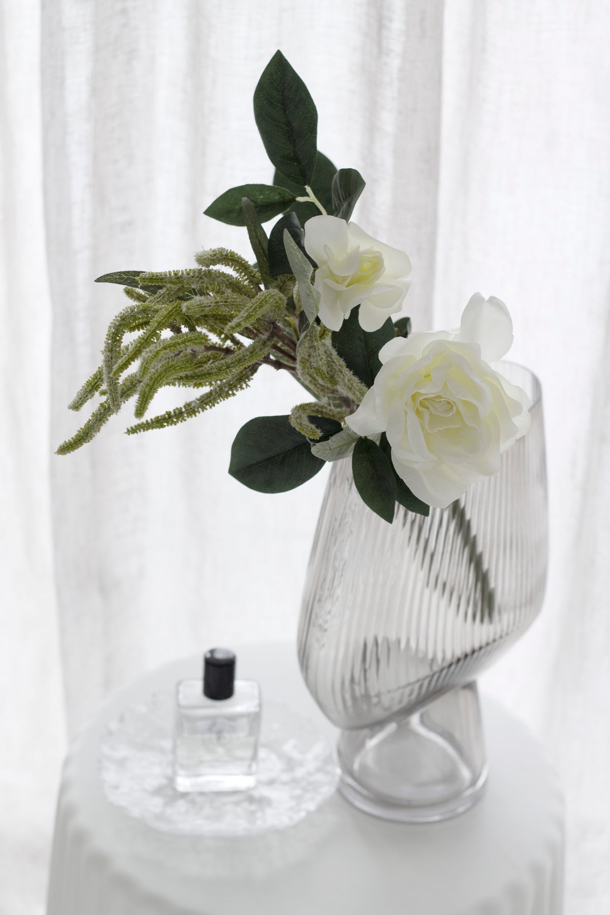 Close-up of a clear ribbed glass vase holding realistic white roses and the green Weeps Spray foliage, styled on a round white pedestal table with a perfume bottle.