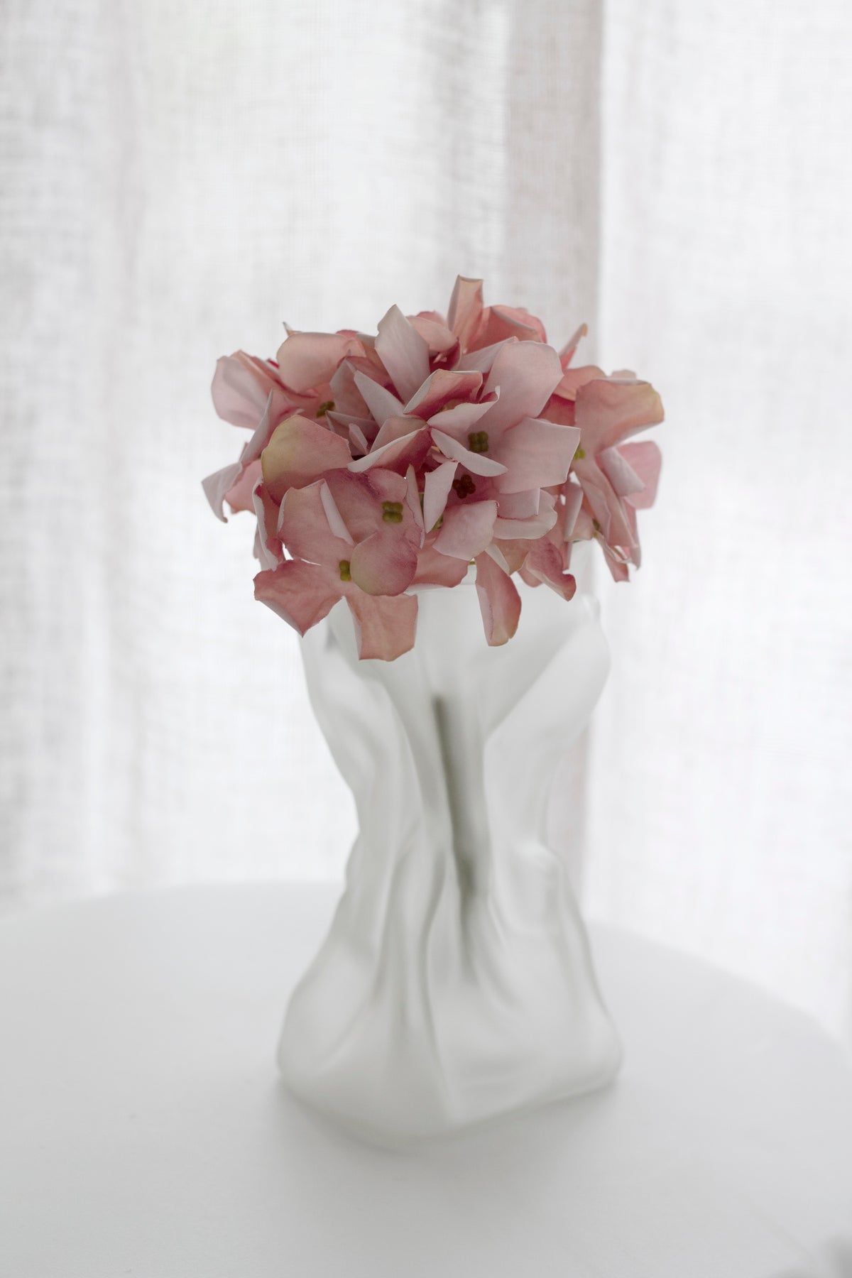Pink faux hydrangea blooms in a white sculpted vase by a curtain