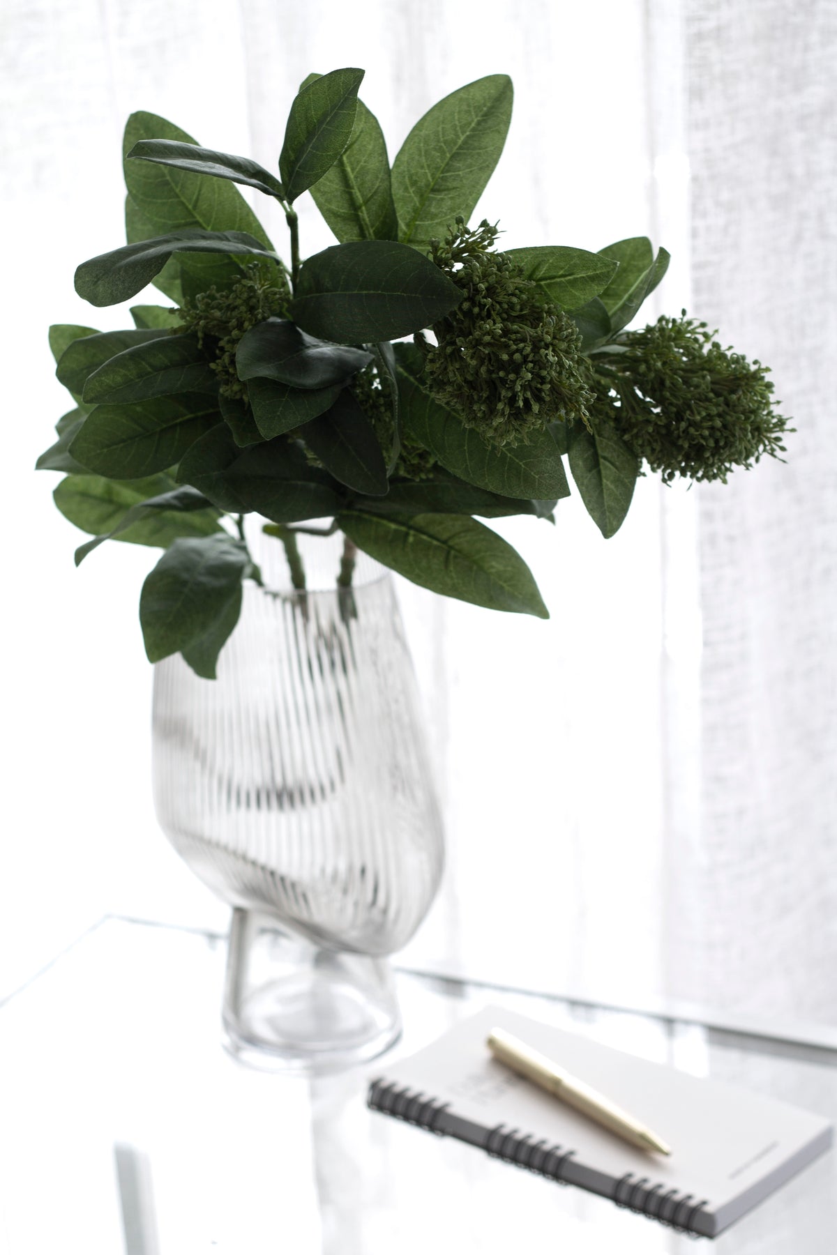 Skimmia in a stemmed ribbed glass vase beside a notebook and pen