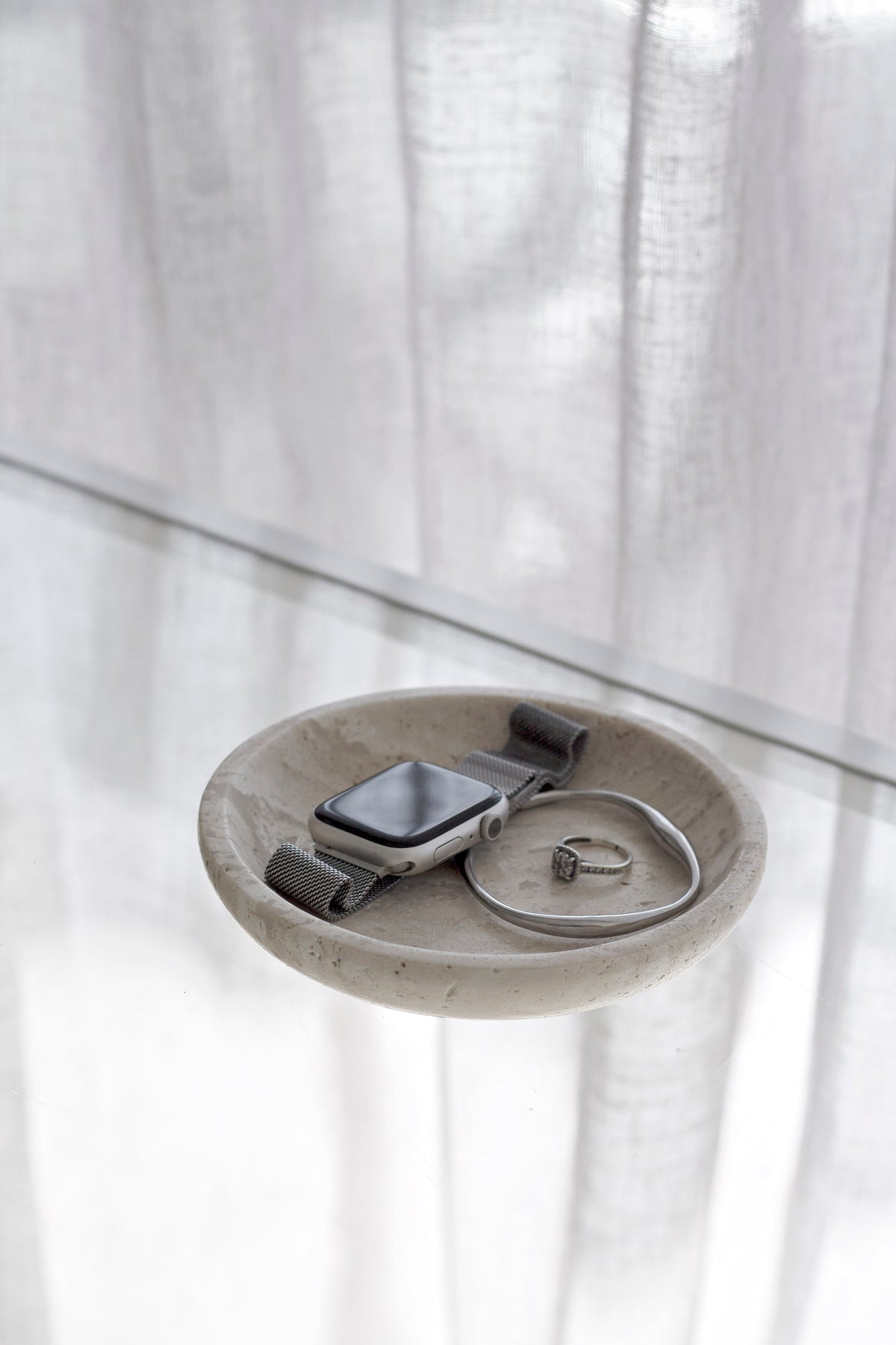 Travertine tray on a clear surface holding a watch and a ring, curtain behind