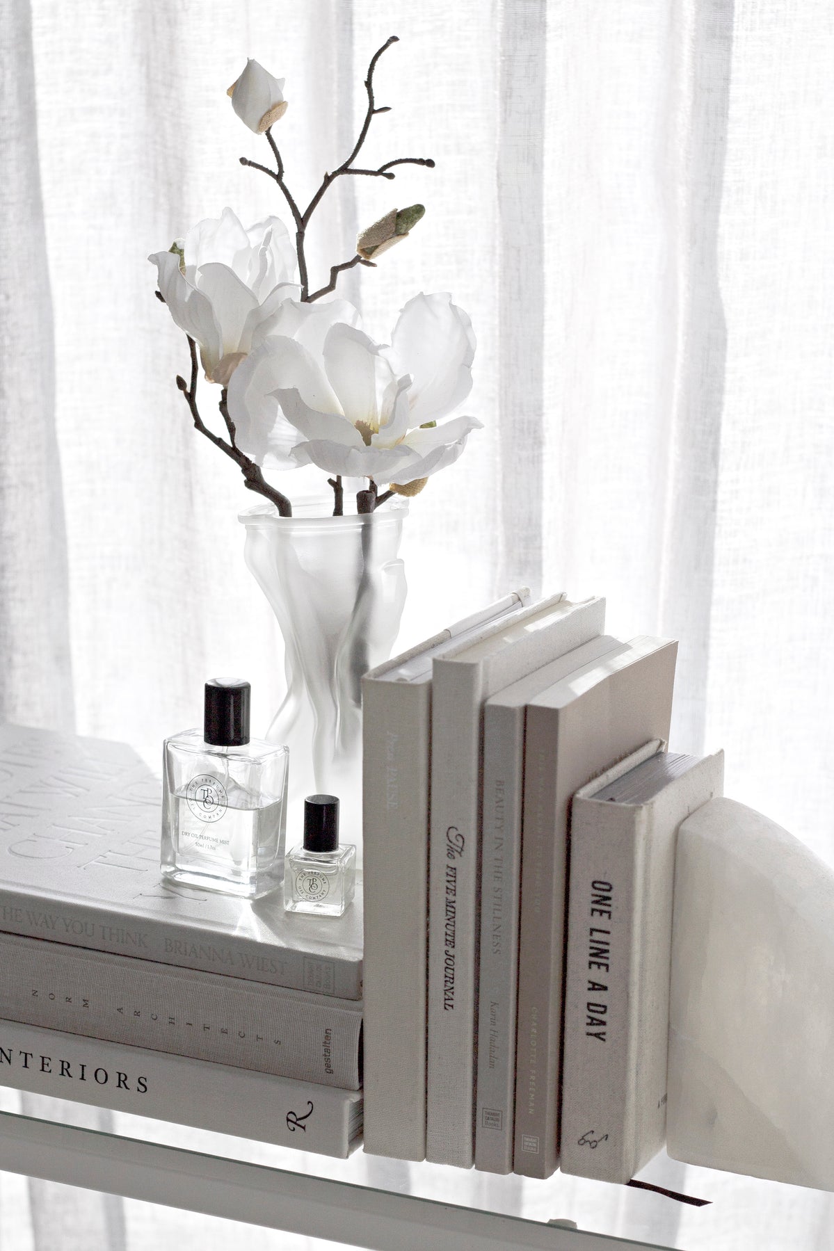 White shelf styled with a vase of blossom, stacked books and small bottles against a sheer curtain