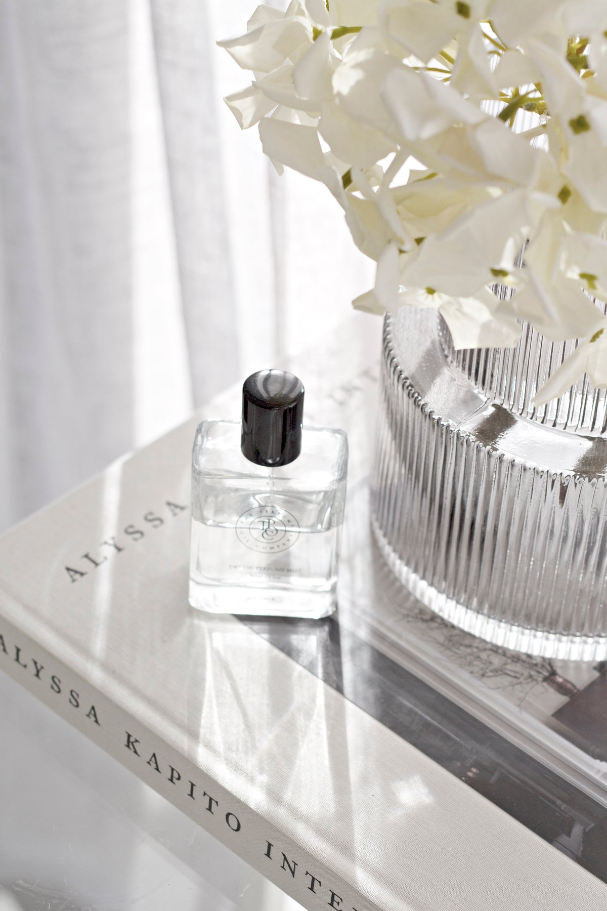 Perfume oil bottle on a marble tray beside a ribbed glass vase of white orchids