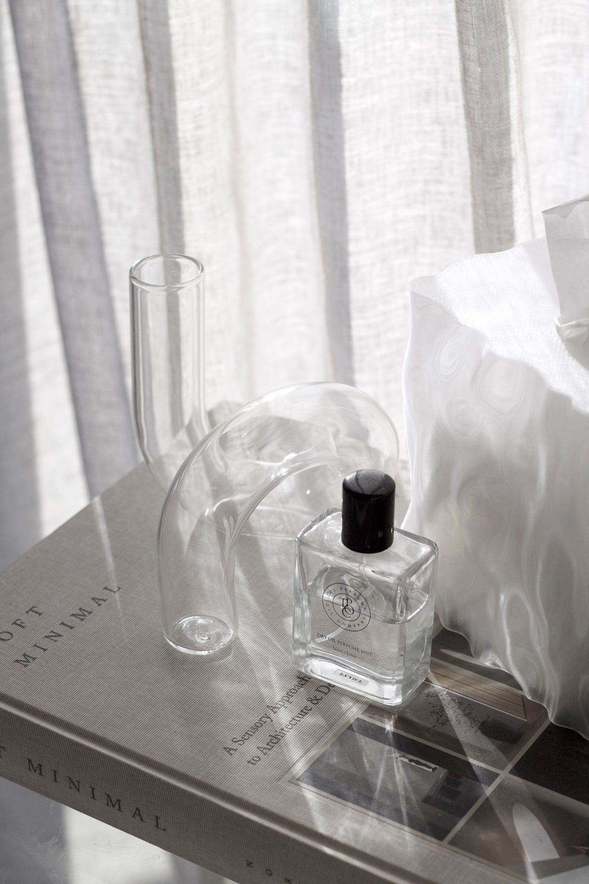 Clear J-Curve vase on books beside a fragrance bottle with light through a linen curtain