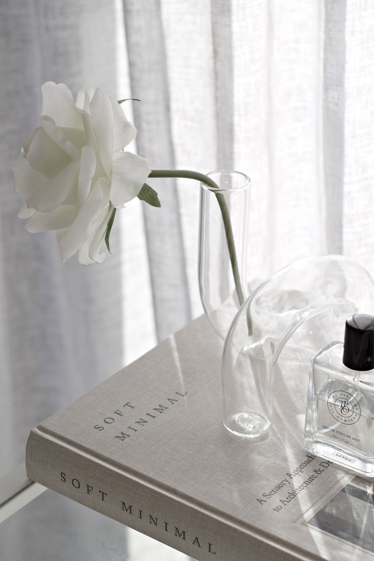 Clear J-Curve vase with a white rose on stacked books beside a small fragrance bottle