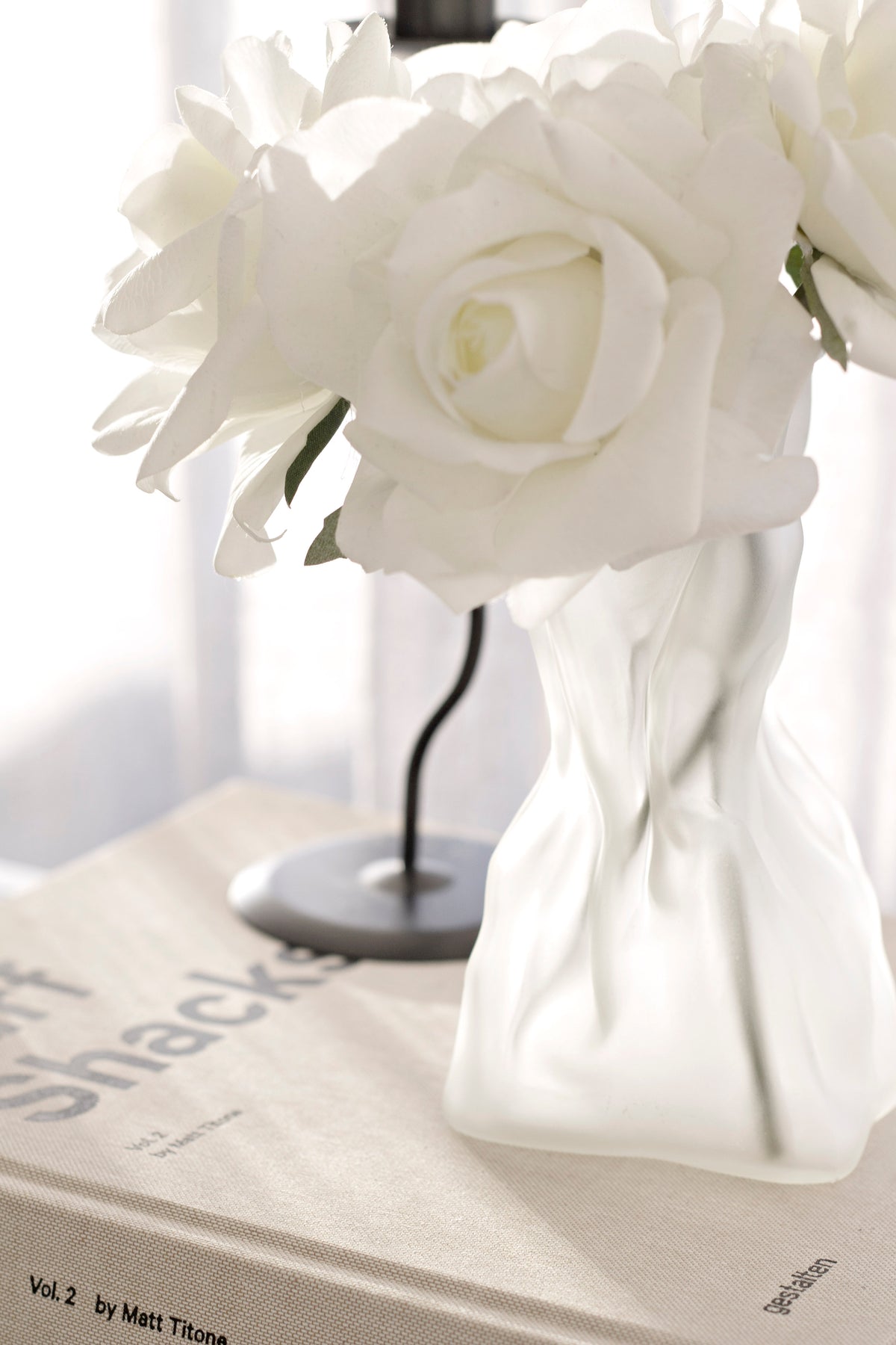 Cream crinkle vase holding white roses on a coffee table book beside a black lamp base