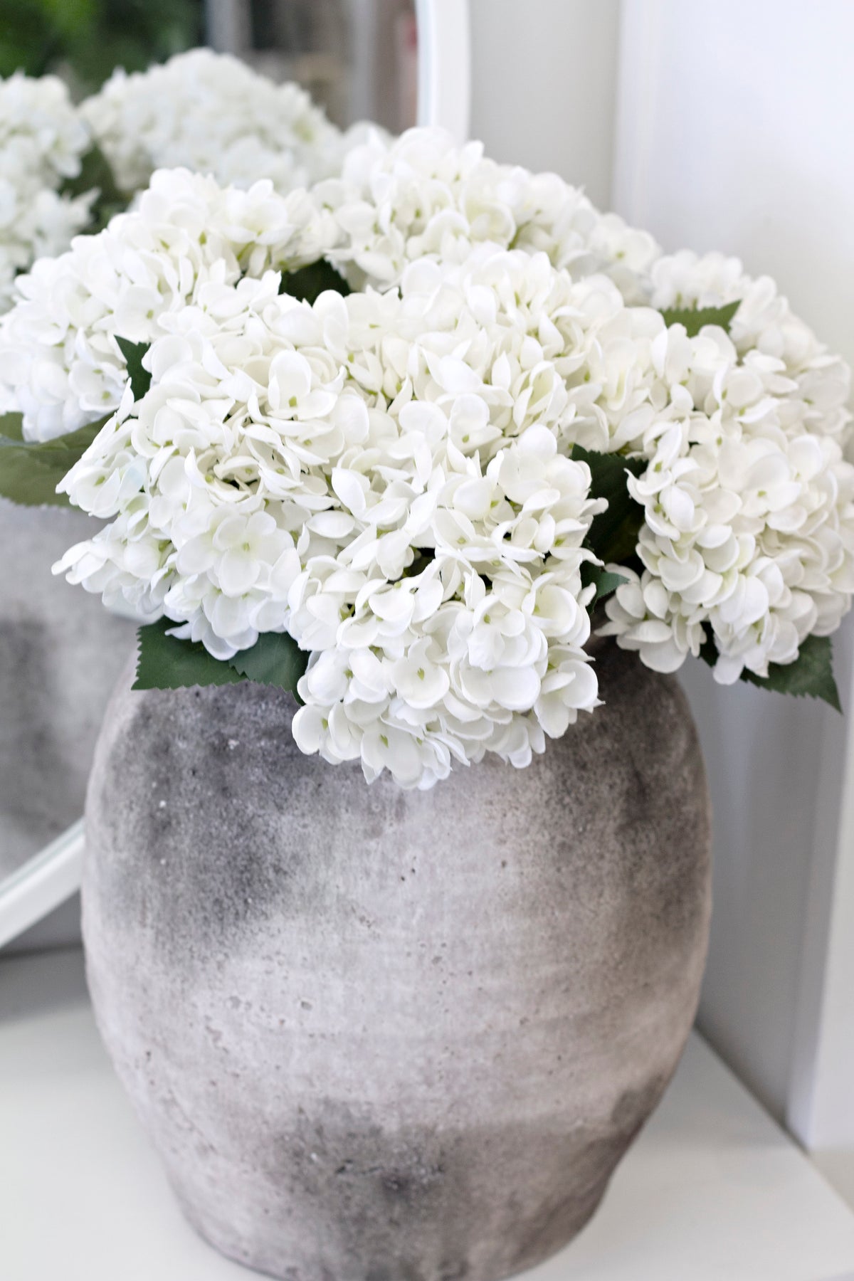 White hydrangeas massed in a mottled grey stone urn with a mirror behind