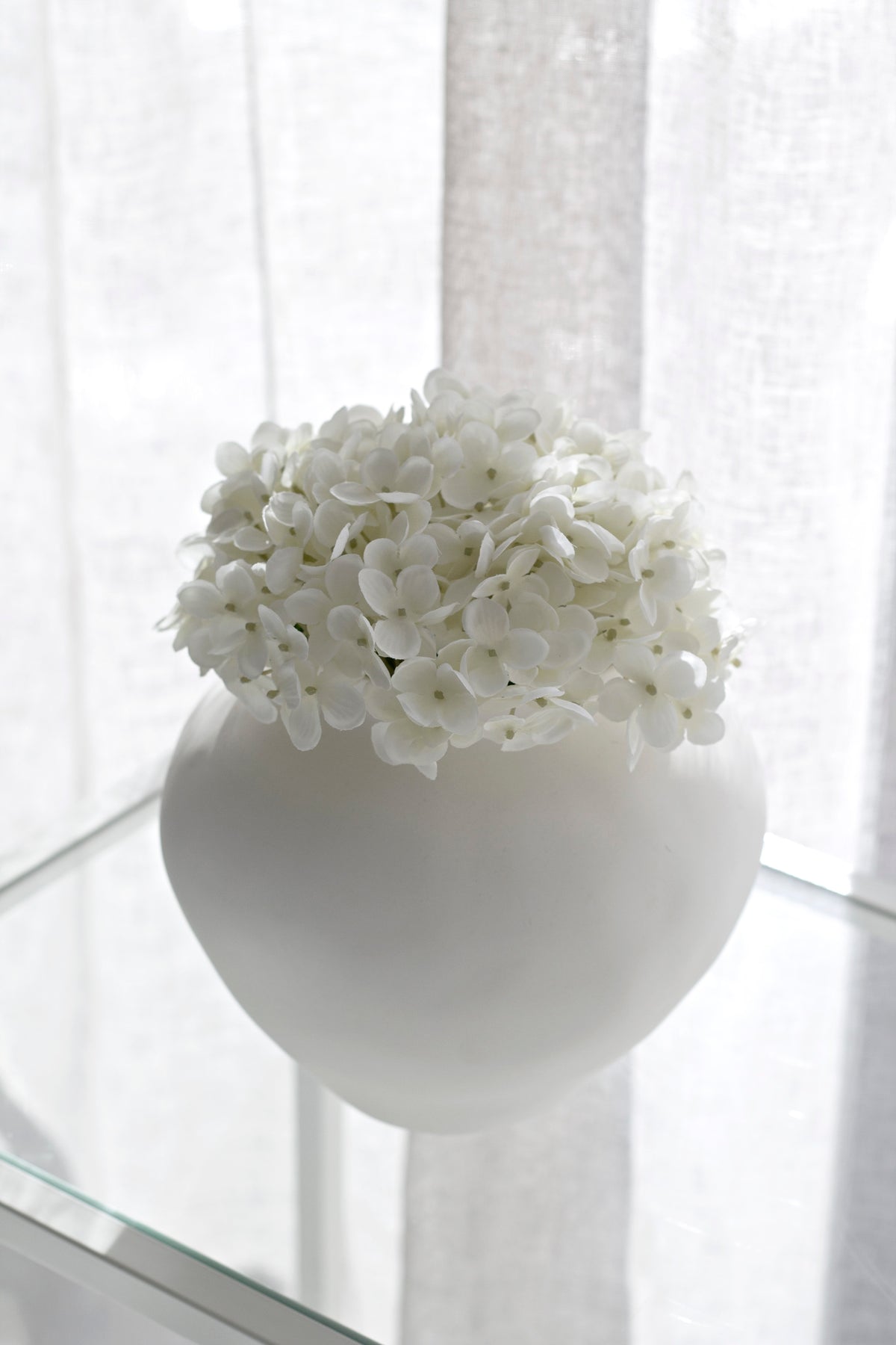 A soft cream faux hydrangea bloom arranged in a white ceramic vase, styled in front of sheer linen curtains on a glass shelf for an airy, elegant look.