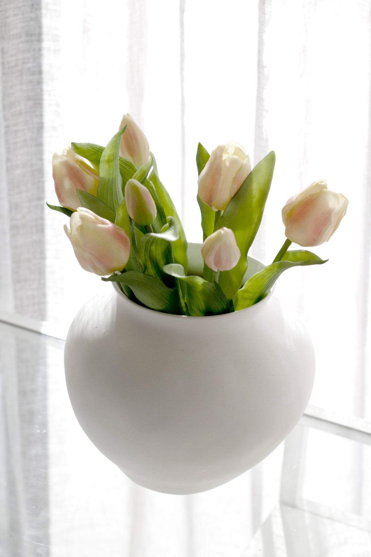 Pale pink faux tulips in a round white vase in front of a sheer curtain