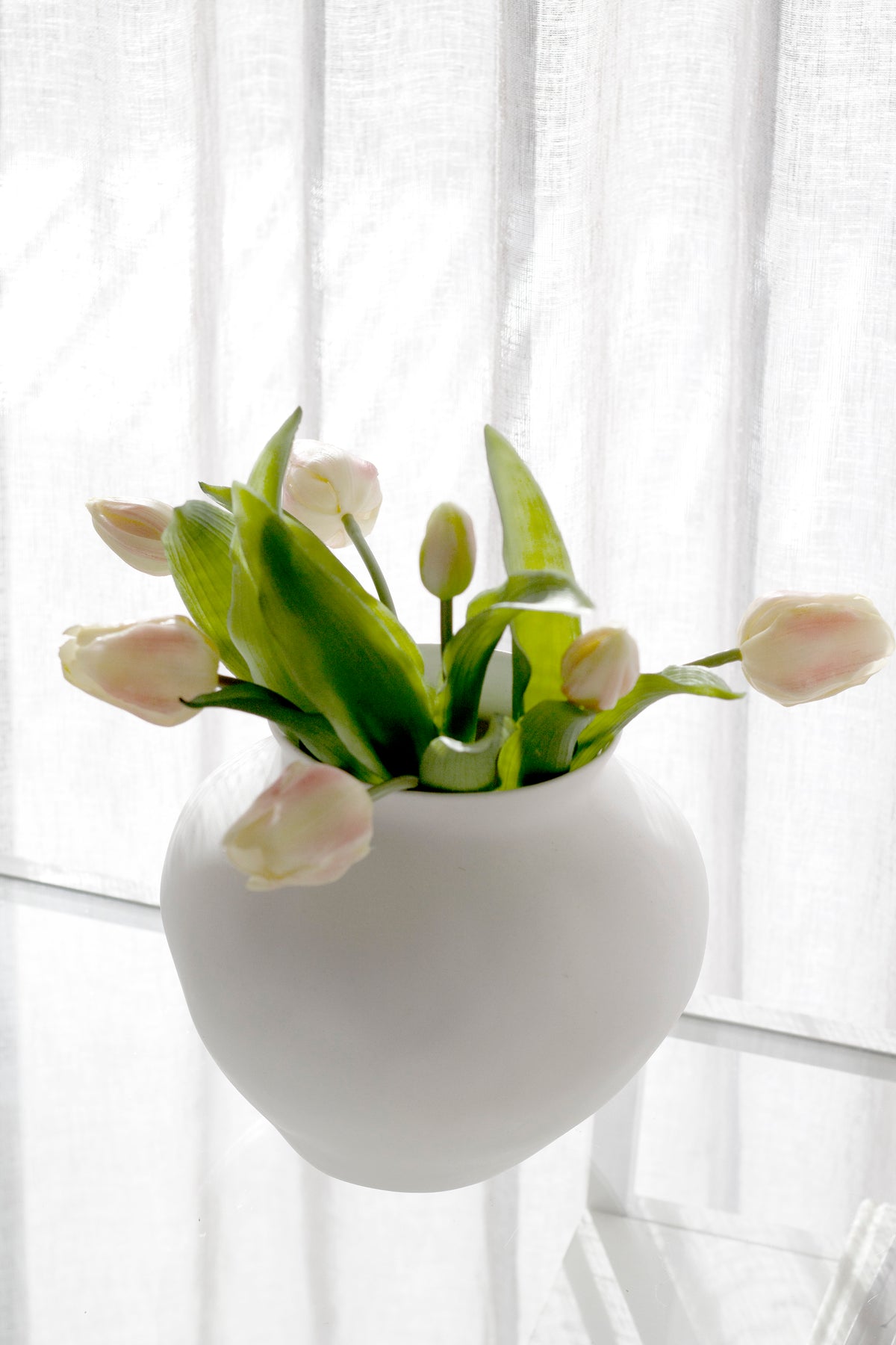 Pale pink faux tulips and buds in a round white vase against a sunlit sheer curtain