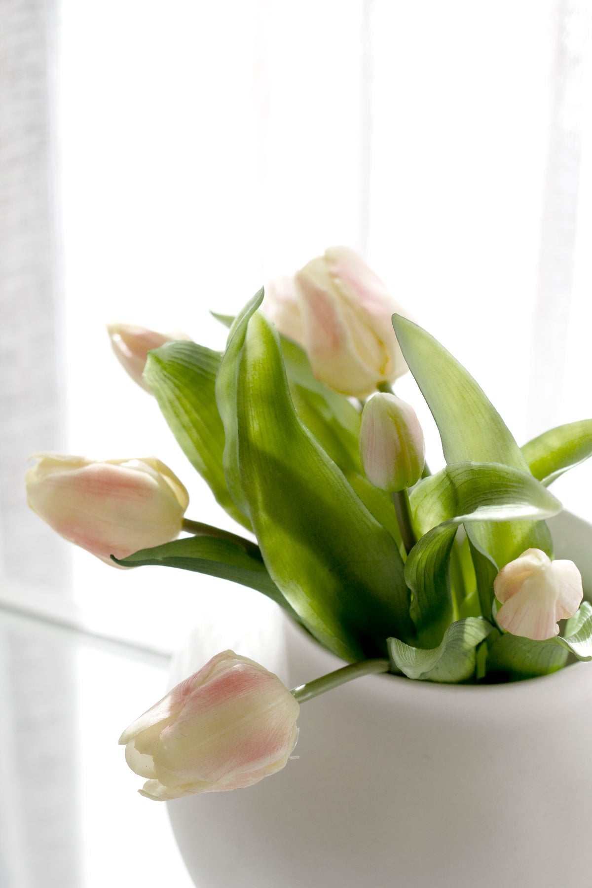 Close up of pale pink faux tulips and buds in a white vase beside a window
