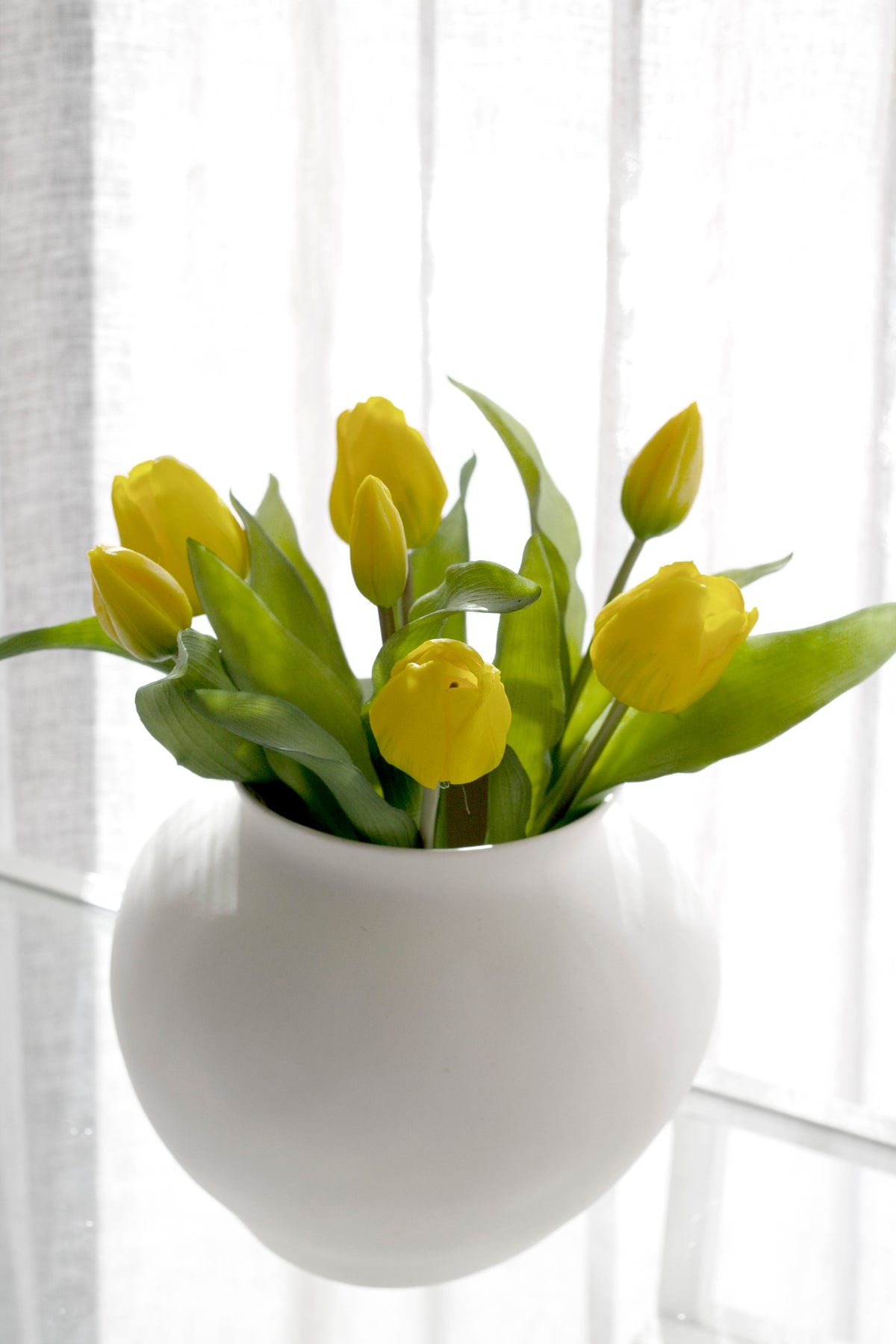 Yellow faux tulips in a round white ceramic vase in front of a sheer curtain