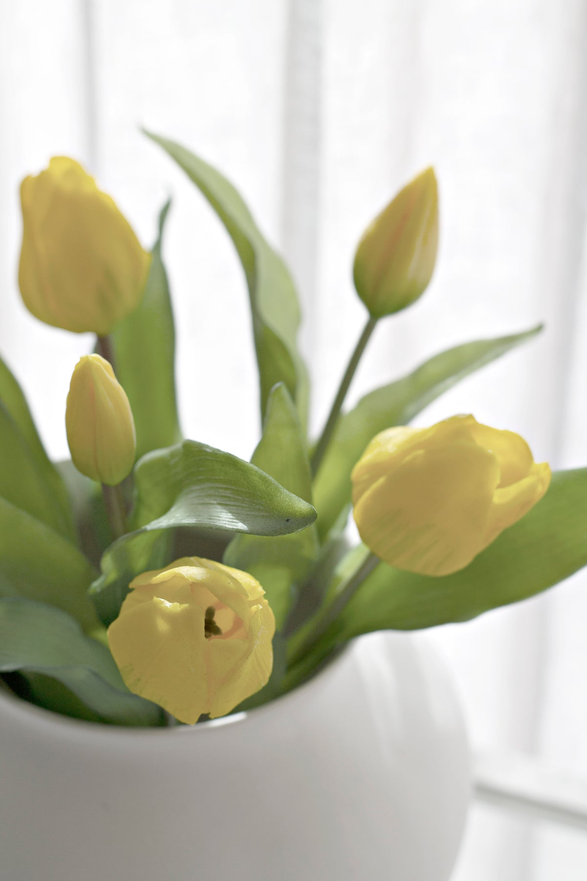 Close up of yellow faux tulips and buds in a white vase against a bright window