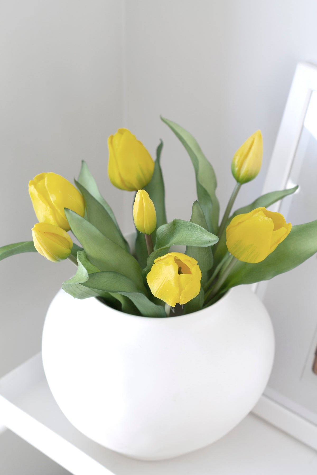 Yellow faux tulips in a round white ceramic vase on a white shelf