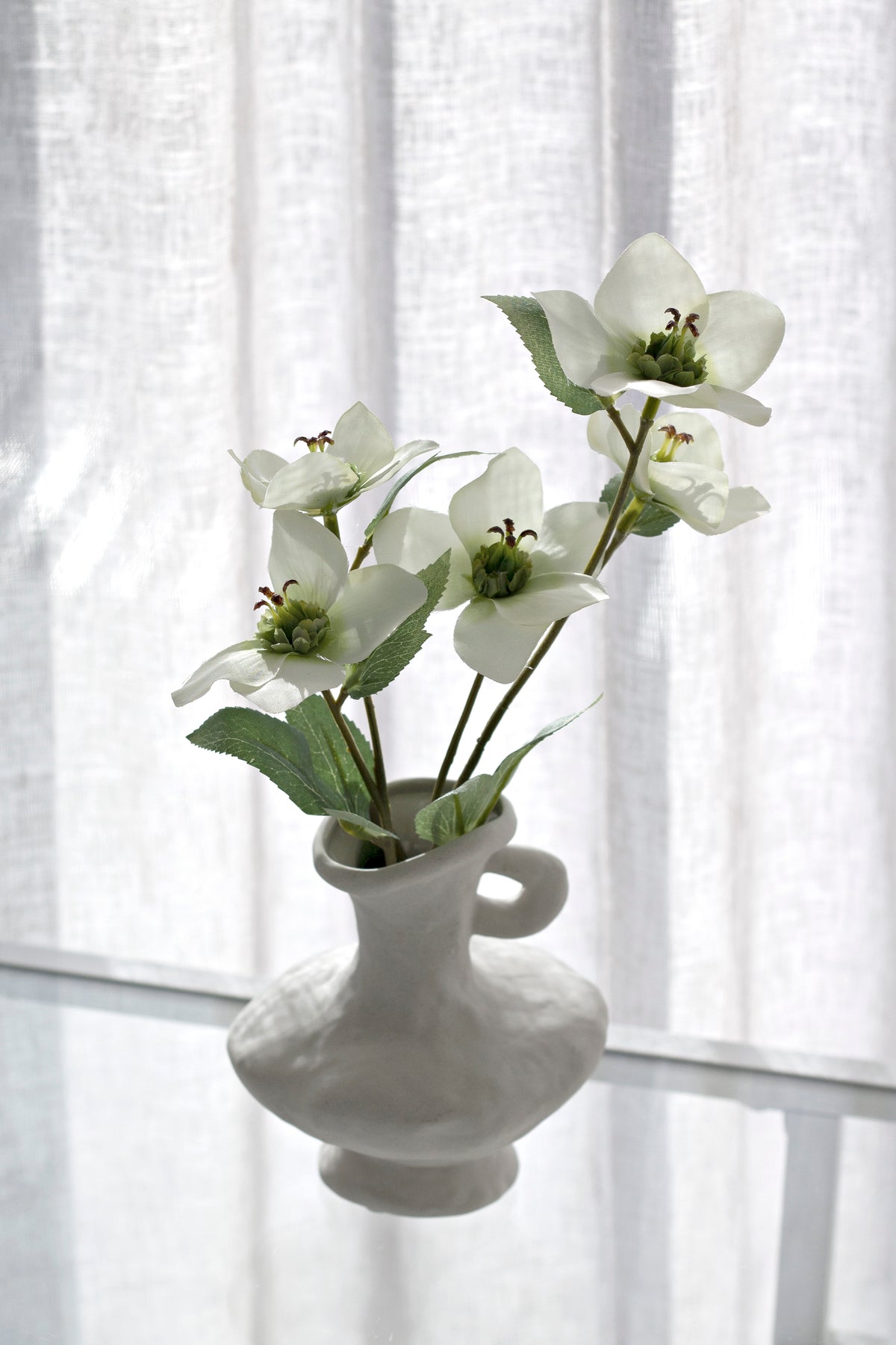 Cream hellebores in a grey handled vase on a glass table by a window